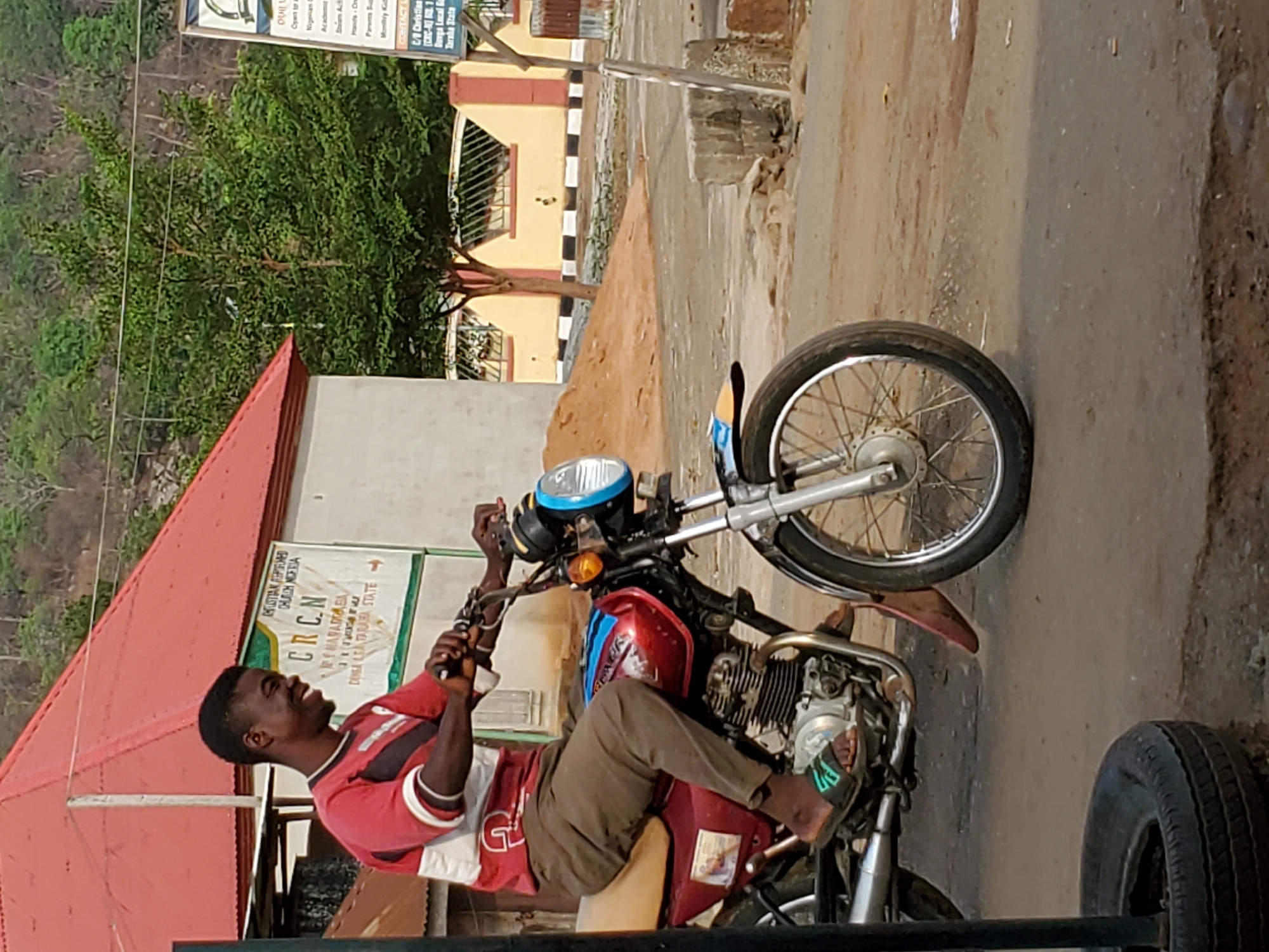 A young man is seated on a red motorcycle, which appears to be part of a modified tricycle or "okada" style transport, indicated by a separate black tire visible in the bottom right foreground. The man, who is Black, is wearing a red and white striped polo shirt, brown trousers, and flip-flops. He is smiling broadly and looking towards his left.
The scene is outdoors during the daytime, with bright sunlight. The motorcycle is positioned on what appears to be an unpaved or dirt road. In the background, to the left, there is a building with a red corrugated metal roof, partially obscured by green trees and distant hills. To the right, another building with yellow walls and black and white striped details (possibly a foundation or decorative element) is visible, with piles of sand or dirt nearby.
A white banner sign is displayed prominently in the background, featuring text related to a religious and educational institution. The legible parts of the text include: "AKUSTIAN ARPITRINED CHURCH NIGERIA", "CRCN", "MARARABA", "DUNGA LEA TARABA STATE", "Nigerian Academy", "Parents Support", and "Monthly Kids Club". This indicates the location is Mararaba, Taraba State, Nigeria. The man's joyful expression suggests a positive or relaxed mood.