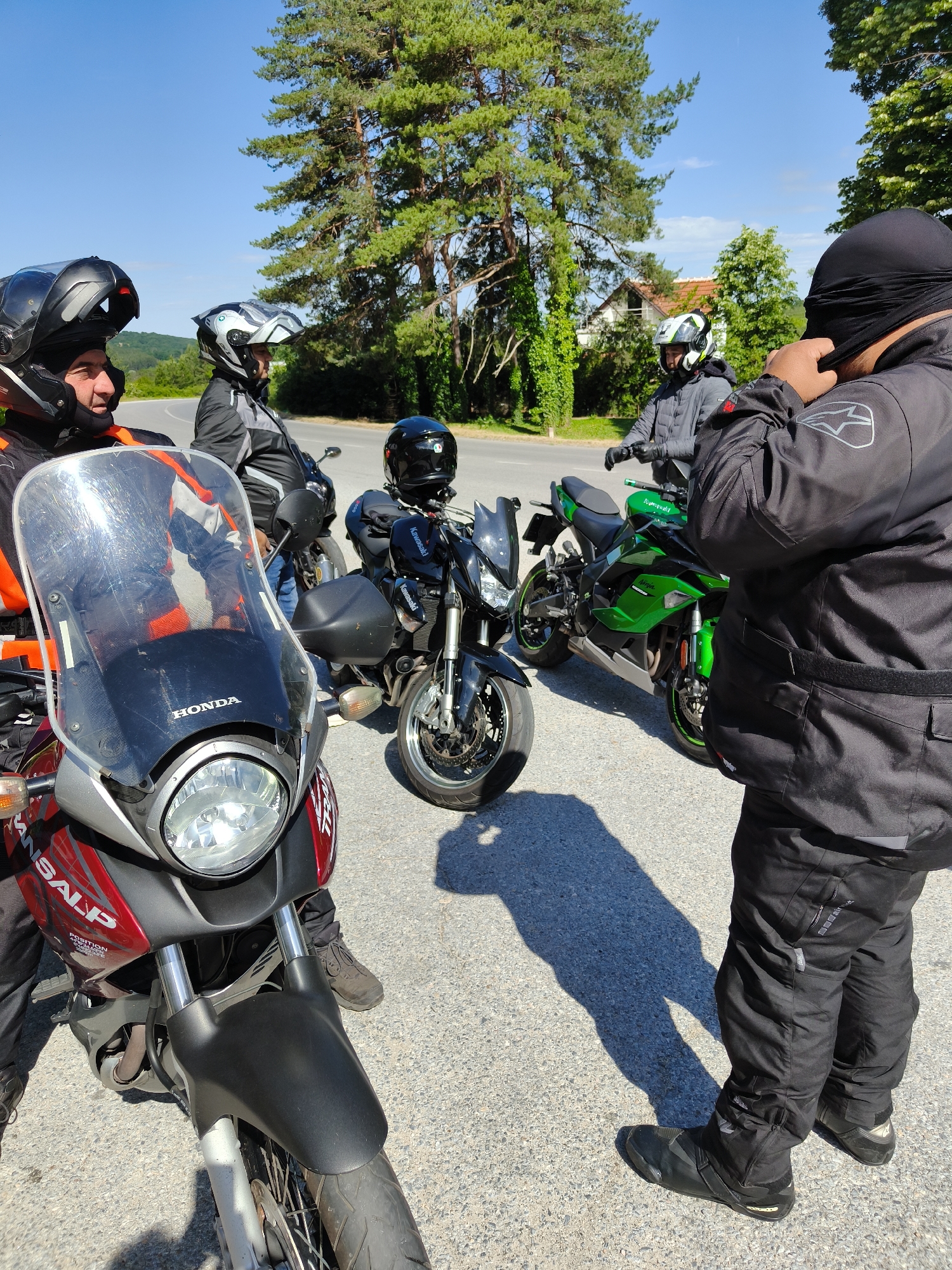 A group of four motorcyclists are gathered on an asphalt road in an unidentified location. The riders are dressed in full motorcycle gear, including helmets and protective clothing. Parked motorcycles include a red Honda Transalp, a black Kawasaki, and a green Kawasaki Ninja. One rider is adjusting his head covering. The environment includes trees, a road, and a distant building. The sky is clear. The scene appears to be a rest stop during a motorcycle ride.