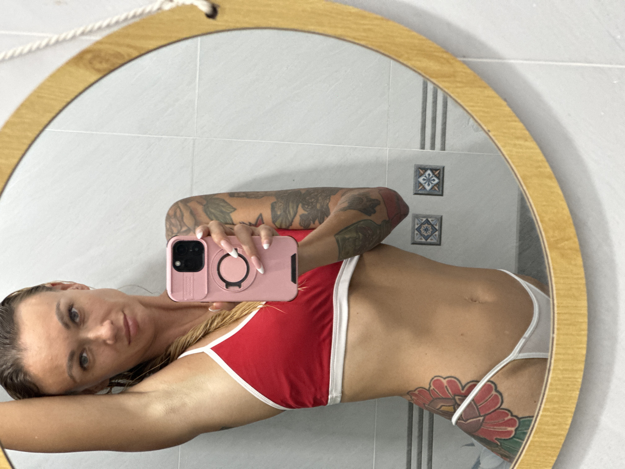 A woman with blonde hair in a braid, wearing a red and white bikini, takes a selfie in a round mirror. Her left arm is raised, and her right hand holds a pink smartphone. She has tattoos on her left arm and hip. The background appears to be a tiled bathroom, with a wooden frame around the mirror. The tiles are white, with some decorative blue and white squares. The overall setting is clean and modern. There is no indication of the location being in Mũi Né, Vietnam.