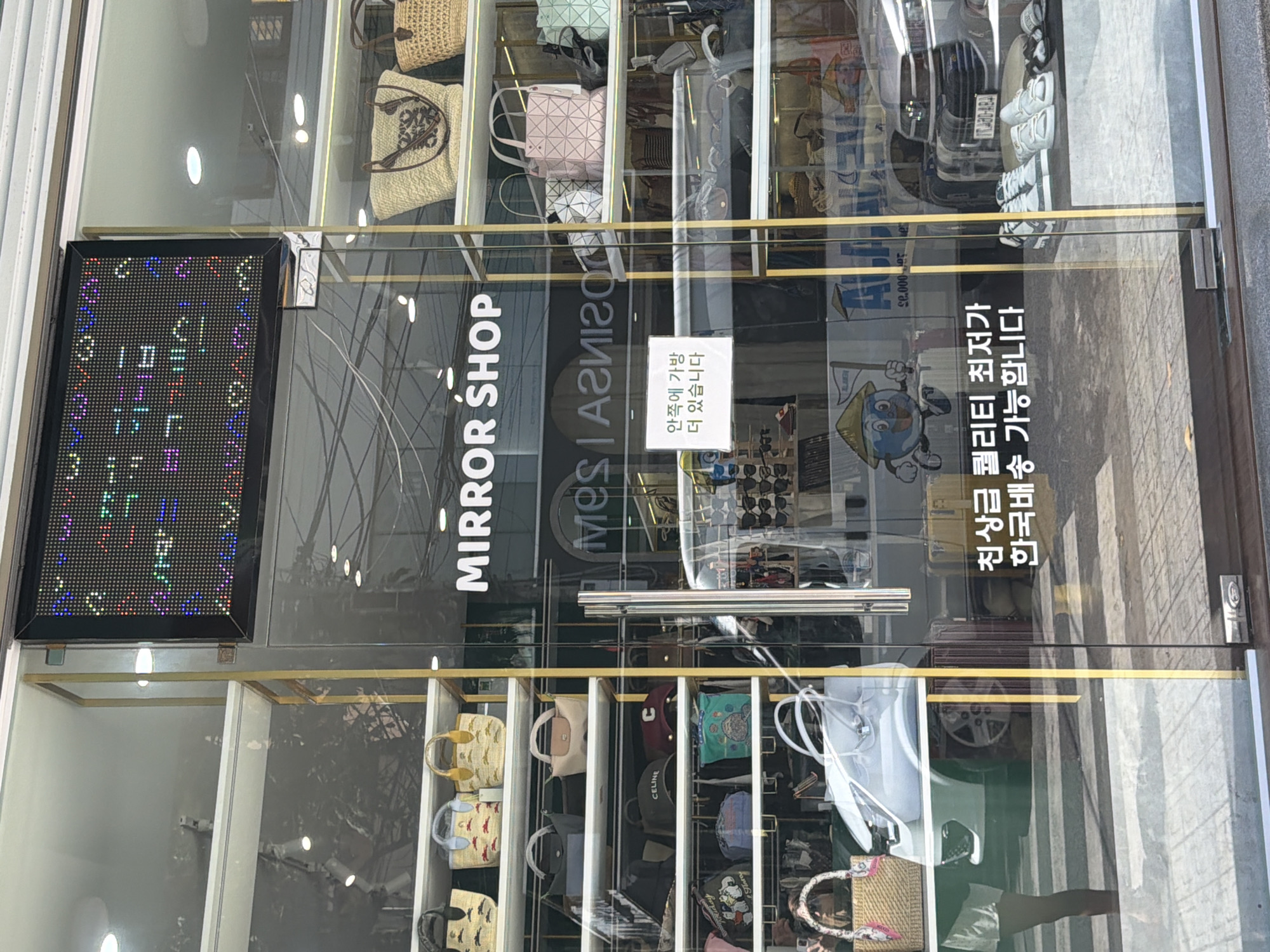 A street-facing shop displays handbags and accessories. The shop, named "MIRROR SHOP", has a glass storefront with shelves stacked with various bags, including straw totes and smaller purses. There's also a white car visible through the glass, and a poster with a cartoon globe character. A sign in Korean indicates "Inside there are more bags". Another sign in Korean reads "Best quality, delivery available". The image is taken during daylight.