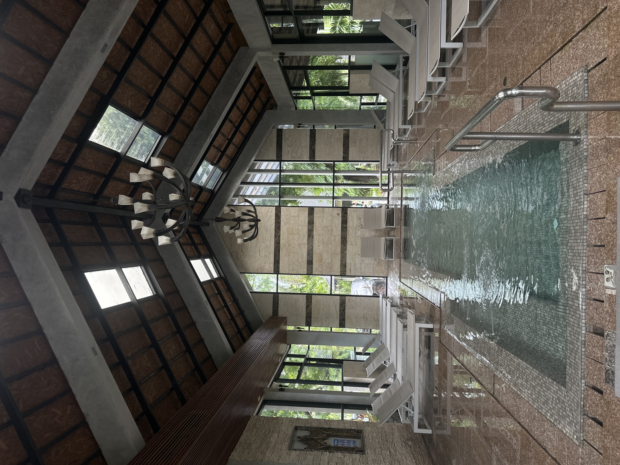 The image depicts an indoor swimming pool area, likely within a spa or hotel. The pool is rectangular with a tiled edge and is surrounded by a polished red-toned floor. Several white lounge chairs are neatly arranged around the pool, with some placed directly on the tiled flooring. The space features a high, vaulted ceiling with exposed wooden beams and translucent skylights that allow natural light to enter. A large, multi-tiered chandelier hangs from the center of the ceiling. Large windows line the walls, offering views of lush green trees and foliage outside, suggesting a natural or tropical setting. The walls are decorated with a combination of stone and patterned tiles. Metal handrails are present near the pool for safety and accessibility. The scene is serene and appears to be unoccupied by people, presenting a tranquil environment.