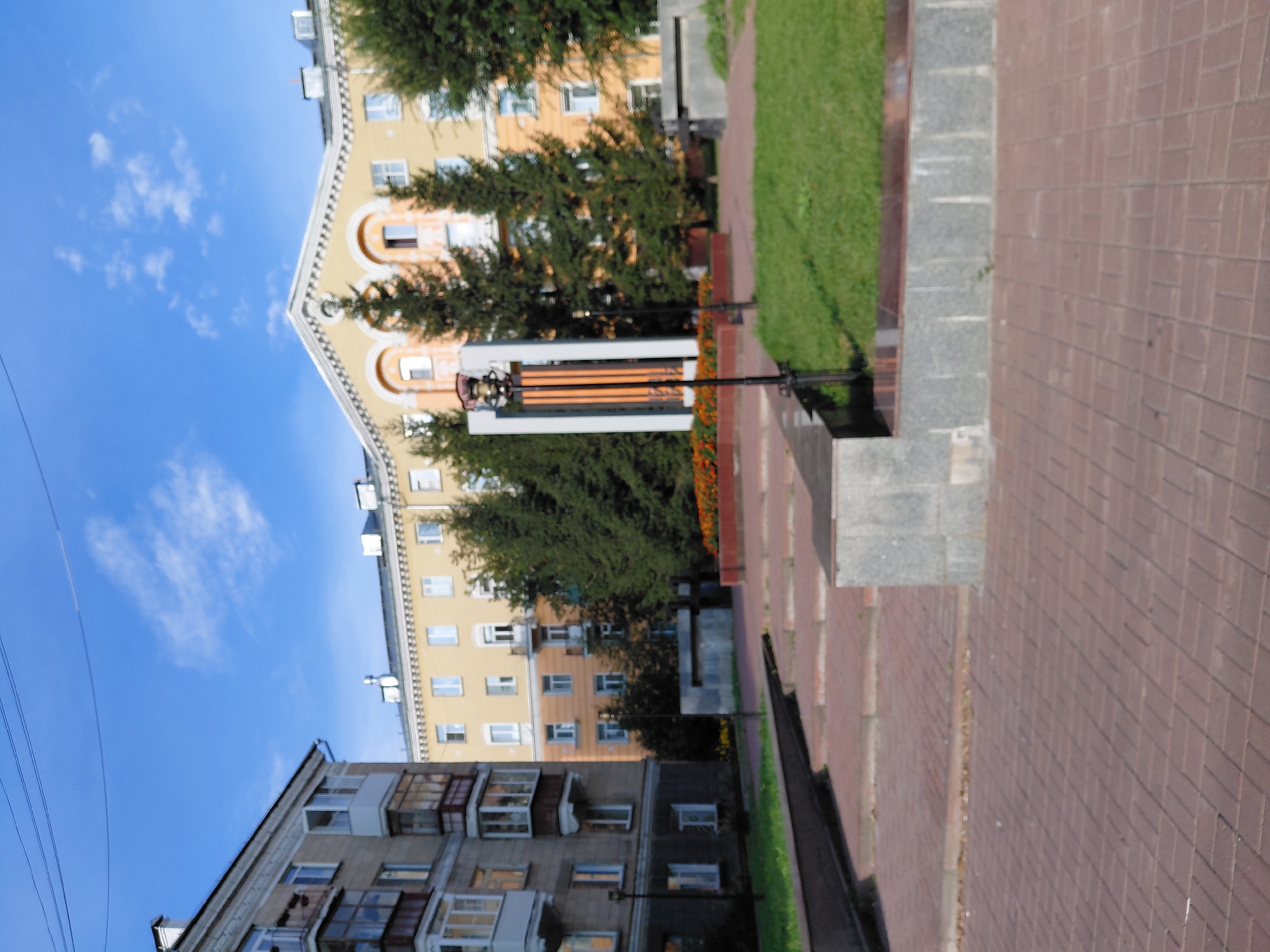 This image shows a view of a courtyard or park area in front of residential buildings in Novosibirsk, Russia. The buildings appear to be Soviet-era apartment blocks with multiple stories, balconies, and a light yellow facade on the larger building. There are trees, green grass, and a paved area with brick-like patterns. A monument or sculpture with vertical elements and orange accents stands in the center of the scene, illuminated by a street lamp. The sky is blue with scattered white clouds. The scene appears to be a public space designed for residents of the surrounding apartment buildings.