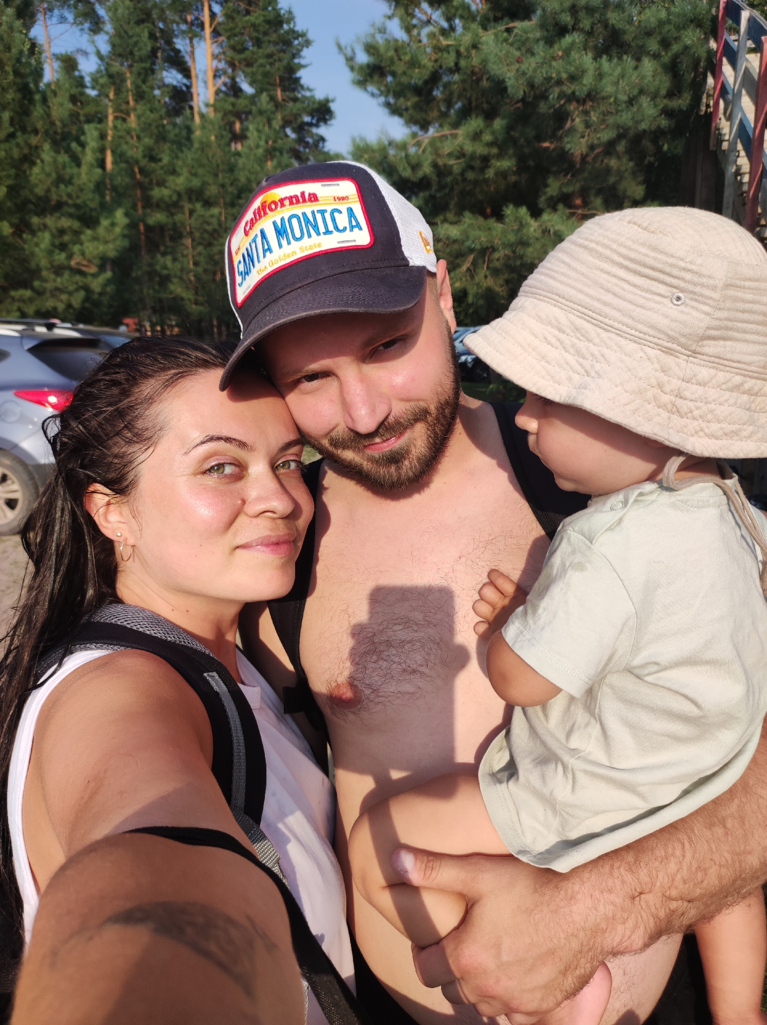 A family, including a man, a woman, and a child, are taking a selfie outdoors. The man is wearing a cap that says "Santa Monica." The woman is next to him, leaning her head on his shoulder. The child is being held by the man. They are in a wooded area with a clear sky. A silver car is visible in the background, as well as a colorful railing structure on the right side of the image. The overall lighting suggests it is daytime, likely late afternoon due to the warm tones and shadows. The location is identified as Novyi, Russia.