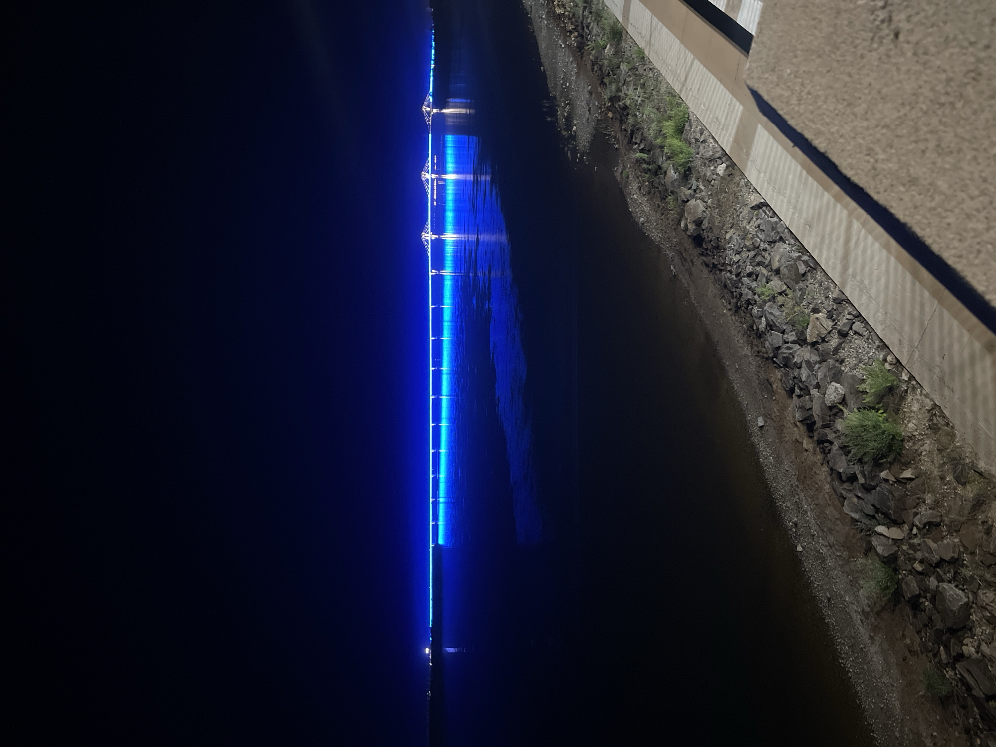 The image depicts the **Amur River Bridge**, also known as the **Blagoveshchensk-Heilongjiang Bridge**, at night. The bridge is illuminated with a vibrant blue light, creating a striking contrast against the dark sky and water. The bridge spans across the Amur River, connecting Blagoveshchensk, Russia, with Heihe, China. The river's surface reflects the bridge's blue illumination, creating a shimmering effect. The foreground of the image shows a rocky embankment and a paved walkway, suggesting a promenade or viewing area. The overall scene conveys a sense of nighttime beauty and tranquility, highlighting the architectural significance of the bridge as a modern landmark.
