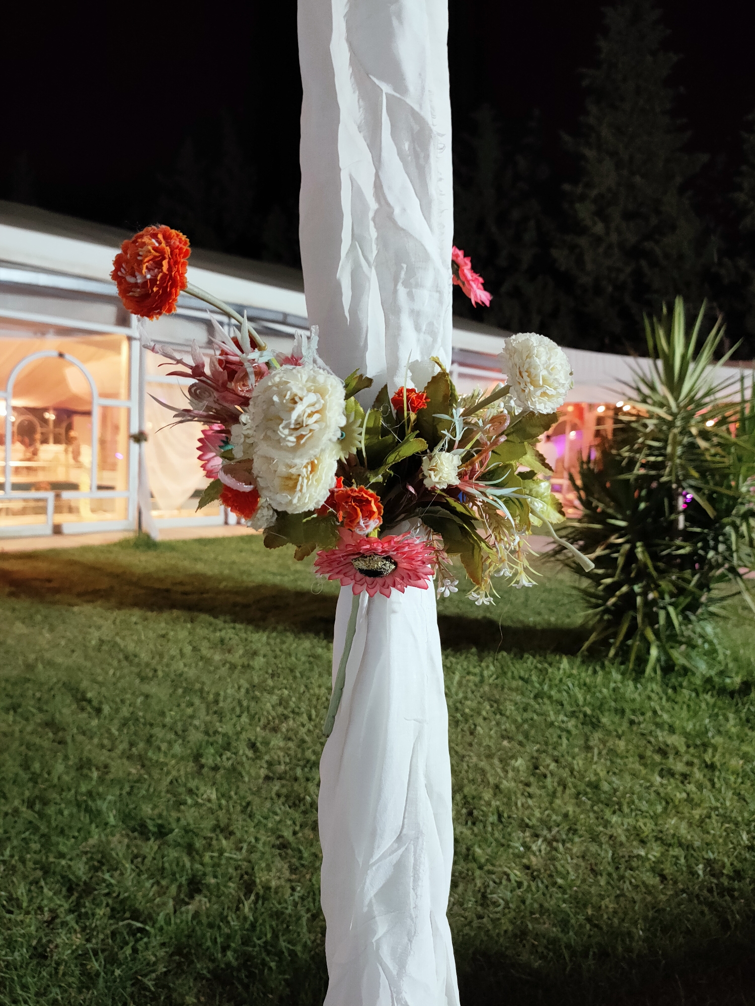 The image shows a floral arrangement attached to a white fabric draped around a vertical pole, outdoors at night. The flowers are a mix of artificial and possibly real, with prominent orange, white, and pink blooms. In the background, a large, lit tent or building with arched entrances is visible, suggesting an event venue. Green grass covers the foreground, and trees are silhouetted against the dark night sky in the distance. A yucca plant stands to the right of the pole. The scene appears to be a decorative element for an outdoor event, possibly a wedding or party, located in the city of Mornag, Tunisia. There are no people visible in the image.