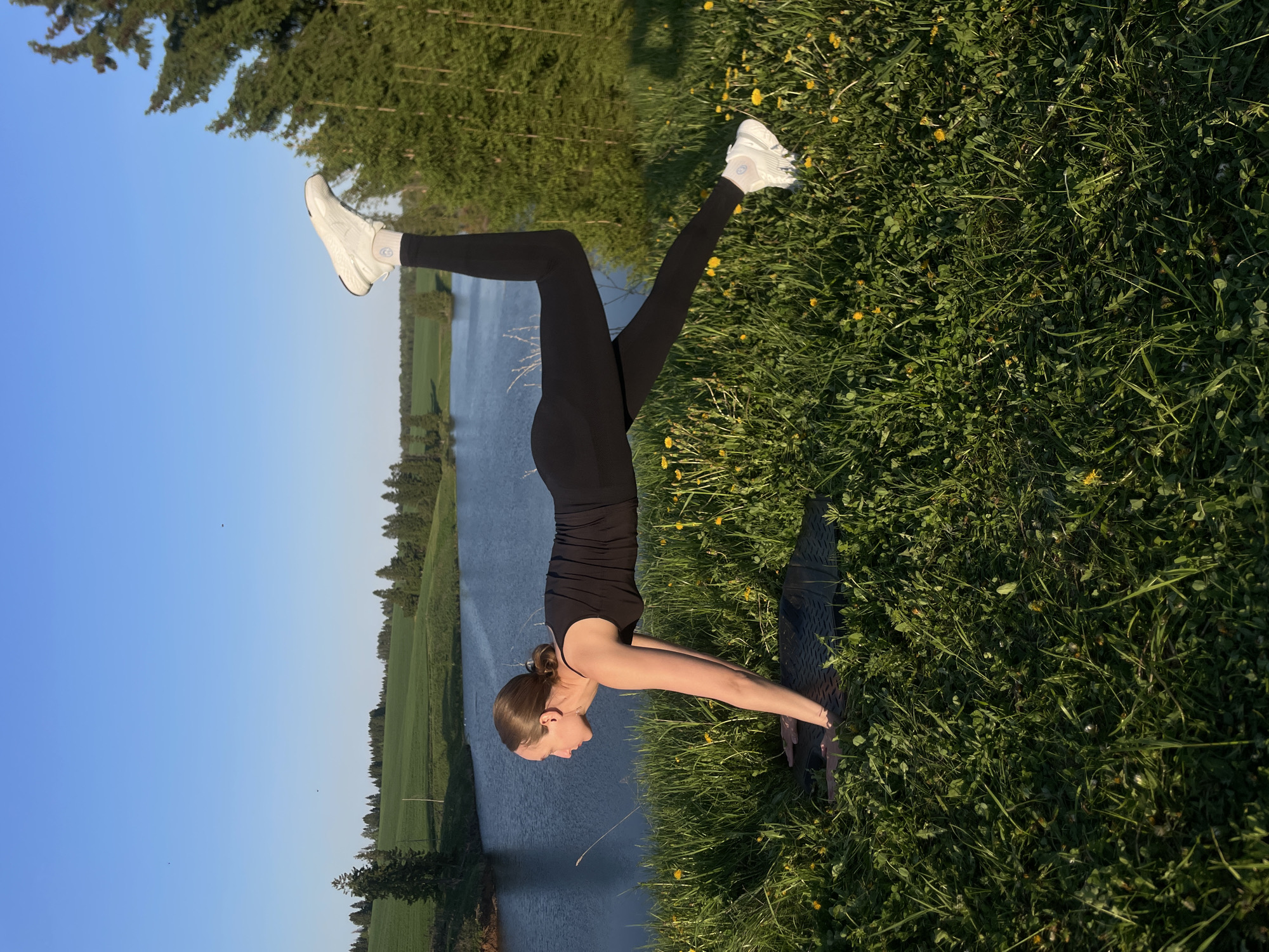 A woman is performing a handstand on a yoga mat in a grassy field with dandelions. She is wearing a black tank top and leggings with white athletic shoes. In the background, there is a body of water, possibly a river or lake, with green fields and trees on the opposite bank. The sky is clear and blue. The photo was taken in Подшивалово, Russia.