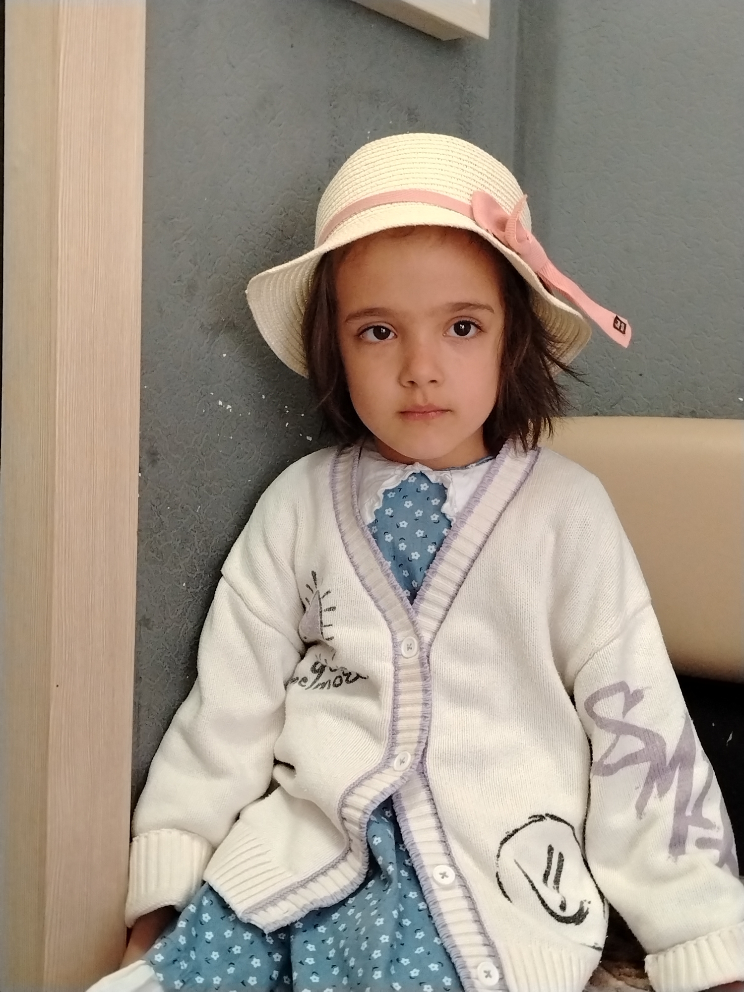 A young girl is sitting indoors against a grey wall, near a light wood-colored door frame. She is wearing a straw hat with a pink ribbon, a blue floral dress with a white collar, and a white cardigan. The cardigan has decorative patterns, including sketched writing and a drawing of a face. The girl is looking directly at the camera. A beige surface, possibly a seat, is visible behind her. There is no indication this photo was taken in Noviy, Russia.
