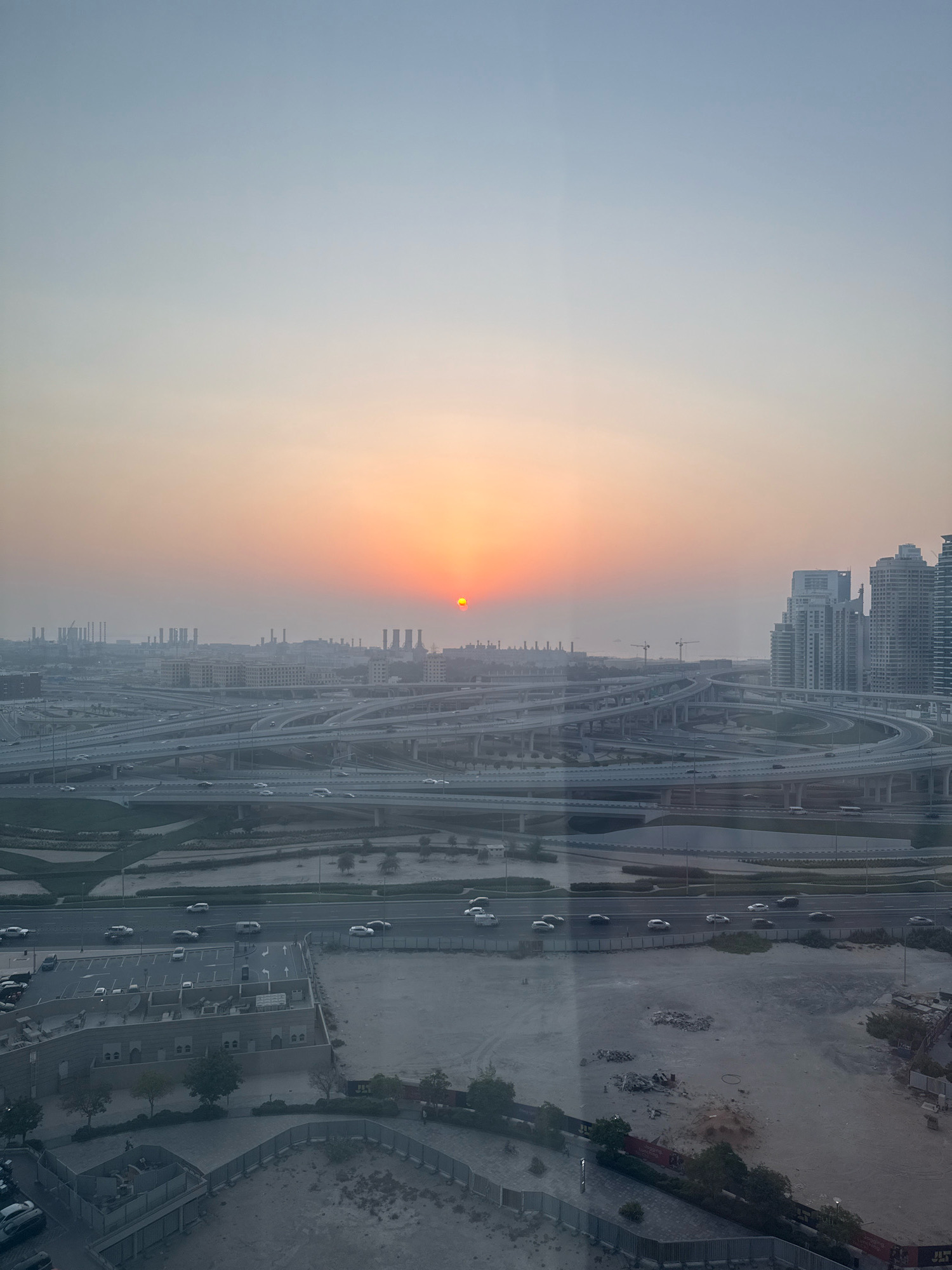 The image depicts a wide-angle view of a sprawling highway interchange in Dubai, United Arab Emirates, at sunset. The sky transitions from a pale blue at the top to a warm orange and pink hue near the horizon, where a small, bright sun is visible. Numerous elevated roads, a signature feature of Dubai's infrastructure, crisscross the scene, forming a complex network. Cars are visible on various lanes of the highway, indicating ongoing traffic. In the background, industrial structures with tall chimneys and modern high-rise buildings, characteristic of Dubai's skyline, are dimly visible through the hazy atmosphere. The foreground shows a construction site or undeveloped land with scattered debris and some greenery. The overall impression is one of a busy, modern metropolis at the end of the day.