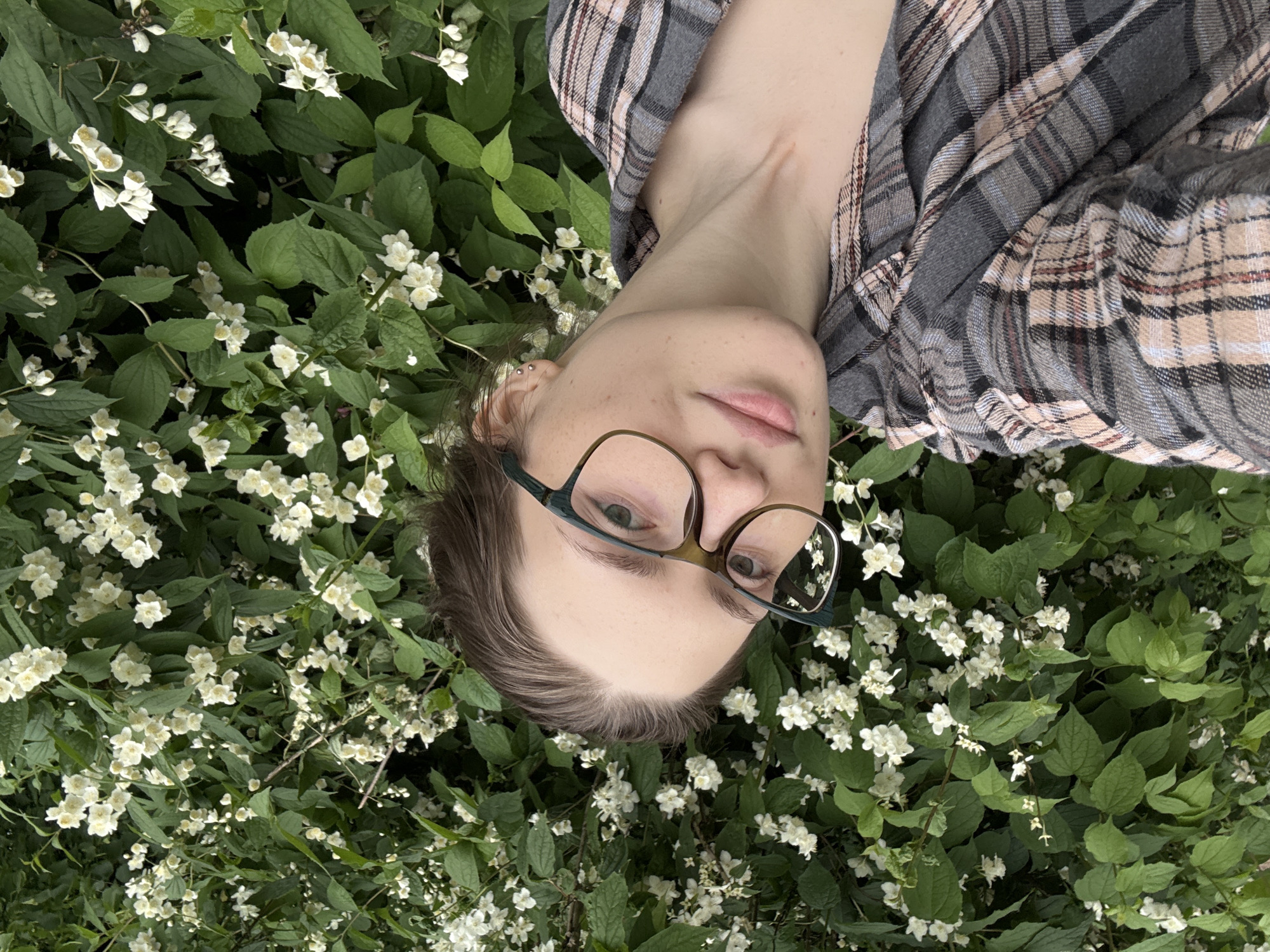 A person with brown hair and glasses is captured in a selfie with a backdrop of green leaves and white flowers. They are wearing a plaid shirt. The image does not contain information indicating a specific location in Saint Petersburg, Russia.