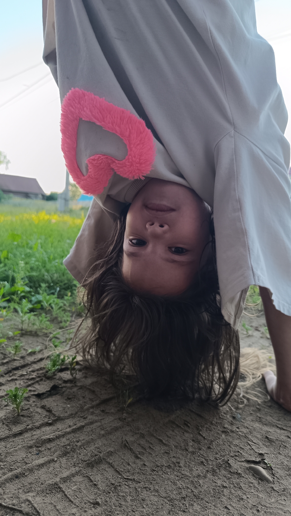 A young girl is pictured upside down in what appears to be a rural setting. She is wearing a light-colored hooded sweatshirt with a pink, fluffy heart-shaped appliqué on the sleeve. Her dark hair is hanging down around her face, obscuring her forehead. Her eyes are open and looking towards the camera. The ground beneath her is dirt with visible tire tracks and small green sprouts. In the background, there is green grass and some yellow wildflowers, with a building and a telephone pole visible in the distance. The sky above is light blue. The image has been cropped and rotated, with the girl's head positioned near the center of the frame. There is no indication of the city of Karsha, Russia.