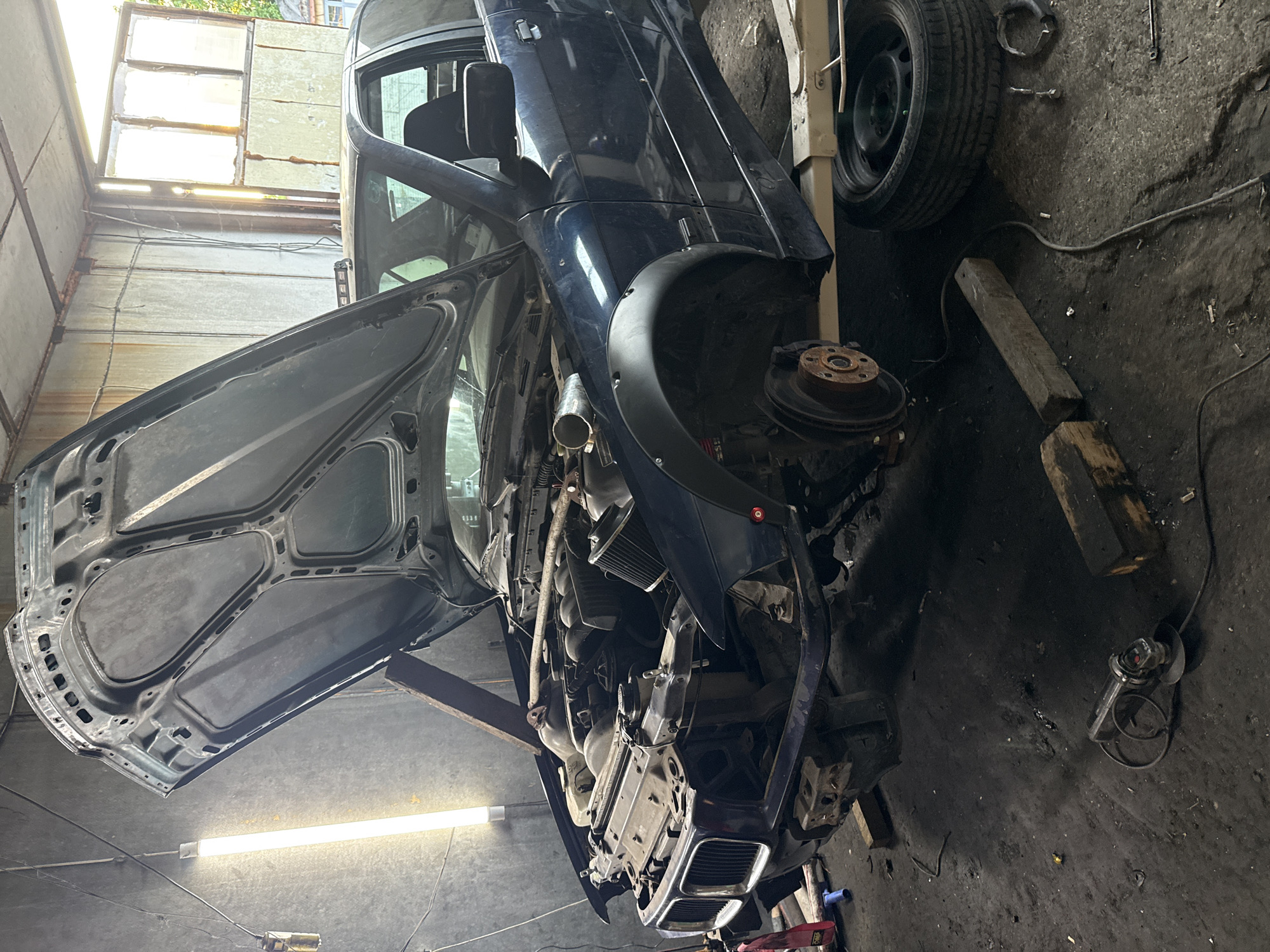 The image depicts an automobile undergoing repairs within a garage setting. The vehicle, a dark-colored passenger car, is elevated, with the hood and driver's side door open, exposing mechanical components. A front wheel has been removed, revealing the brake rotor. Spare tires, tools, and wooden supports are scattered around the garage floor. The walls of the garage are weathered, with exposed brick and patched surfaces. Ambient light enters through a window in the background. Shumen, Bulgaria is not visually verifiable from the image.