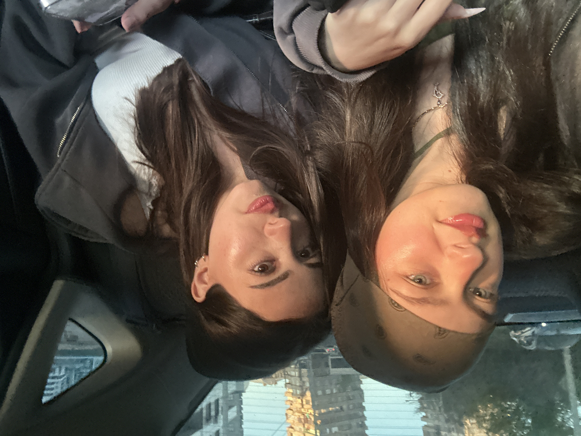 Two women are pictured inside a car in Yerevan, Armenia. One woman wears a white top and gray jacket, while the other wears a bandana. Buildings are visible through the car's windows.