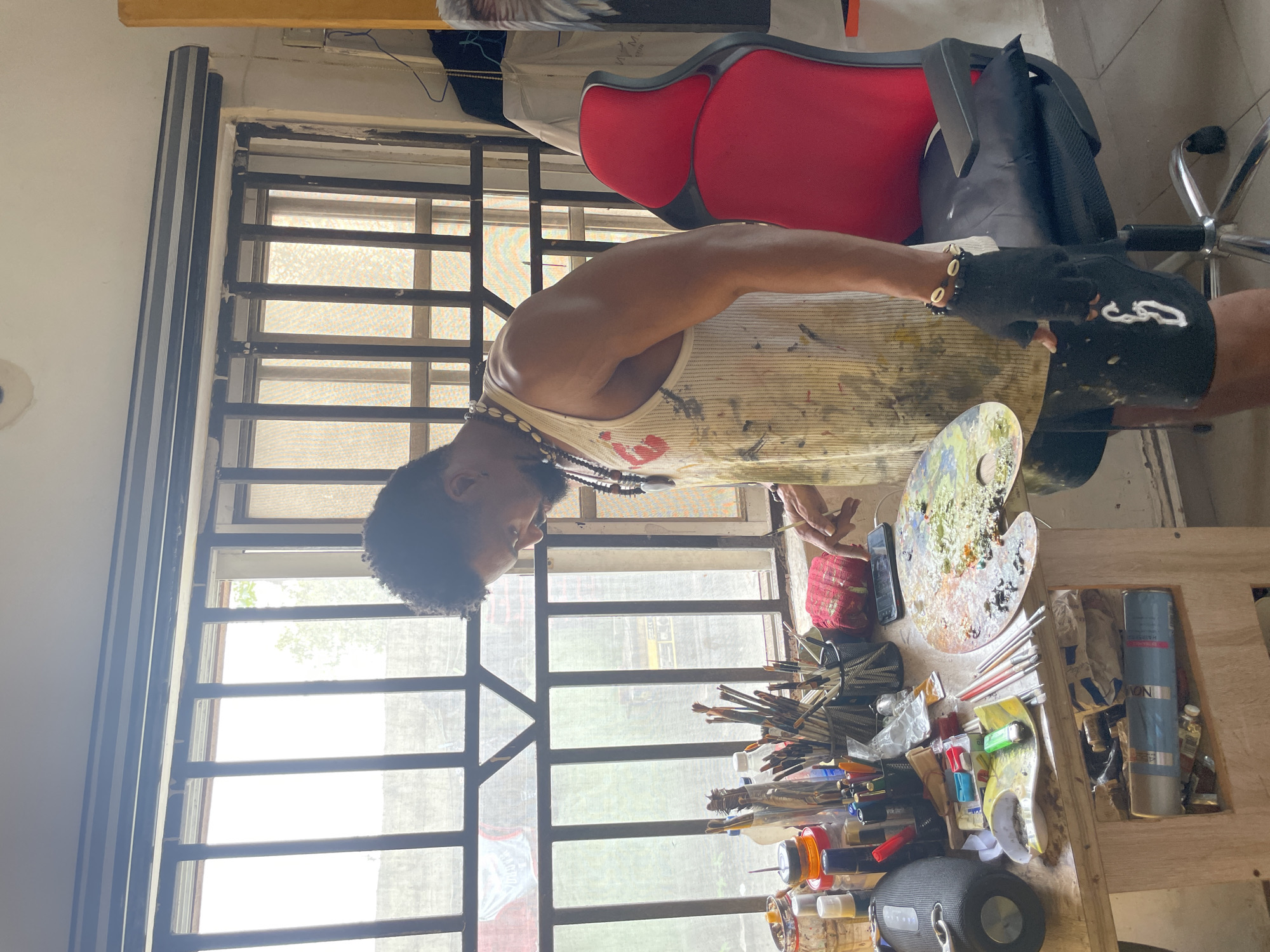 The image depicts a male artist in a well-lit studio, likely located in Ado, Nigeria. He is standing beside a wooden workbench covered with an array of art supplies, including paint tubes, brushes, a palette laden with colorful paints, and various containers. The artist, wearing a paint-splattered tank top and shorts, is focused on his palette, with one hand holding a brush. A red office chair is positioned to his right, and a window with metal bars, partially obscured by curtains, forms the backdrop. The overall environment suggests a working artist's space, with a focus on the creative process.