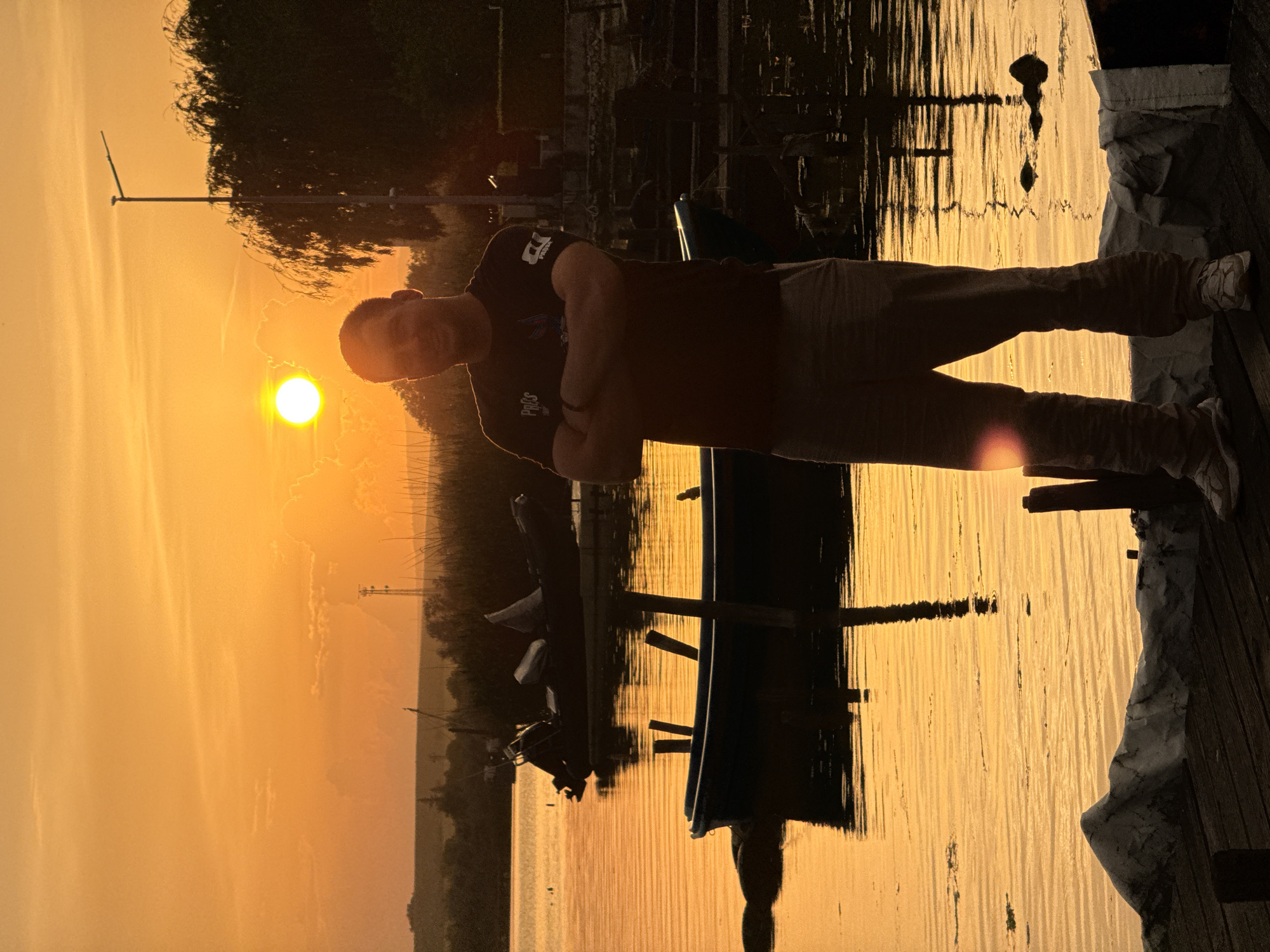 A silhouette of a man stands on a dock by a lake. The location is Казашко, Bulgaria. The sun is setting on the horizon, casting an orange glow on the lake and sky. A small boat sits docked nearby. The man has his arms crossed.