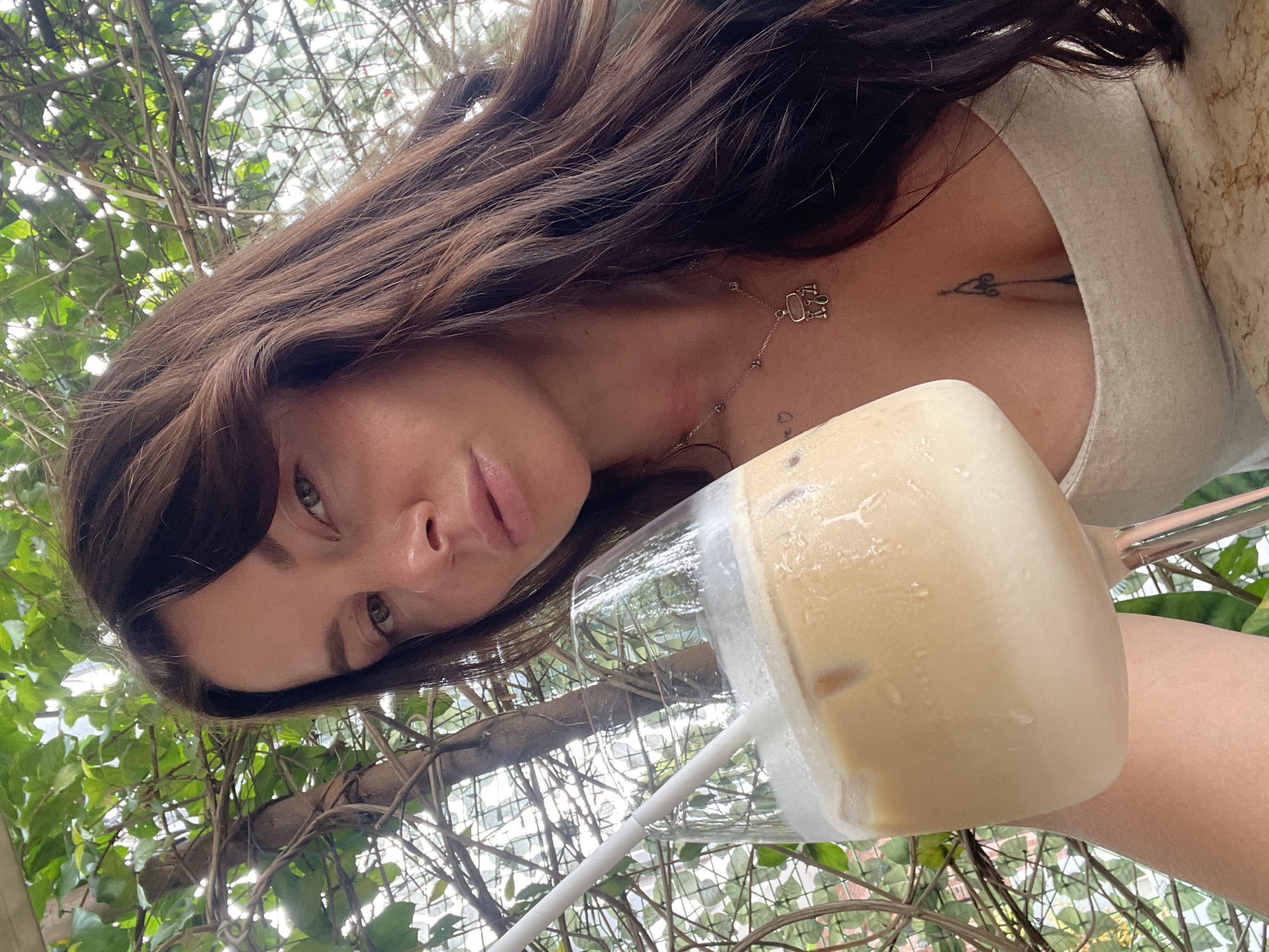 A woman with long, dark brown hair is holding a large glass of iced coffee. She is wearing a light-colored tank top and a necklace. A tattoo is visible on her chest. She is outdoors, with green foliage and branches in the background. The environment suggests a relaxed outdoor setting, possibly a cafe or restaurant in North Kuta, Indonesia.