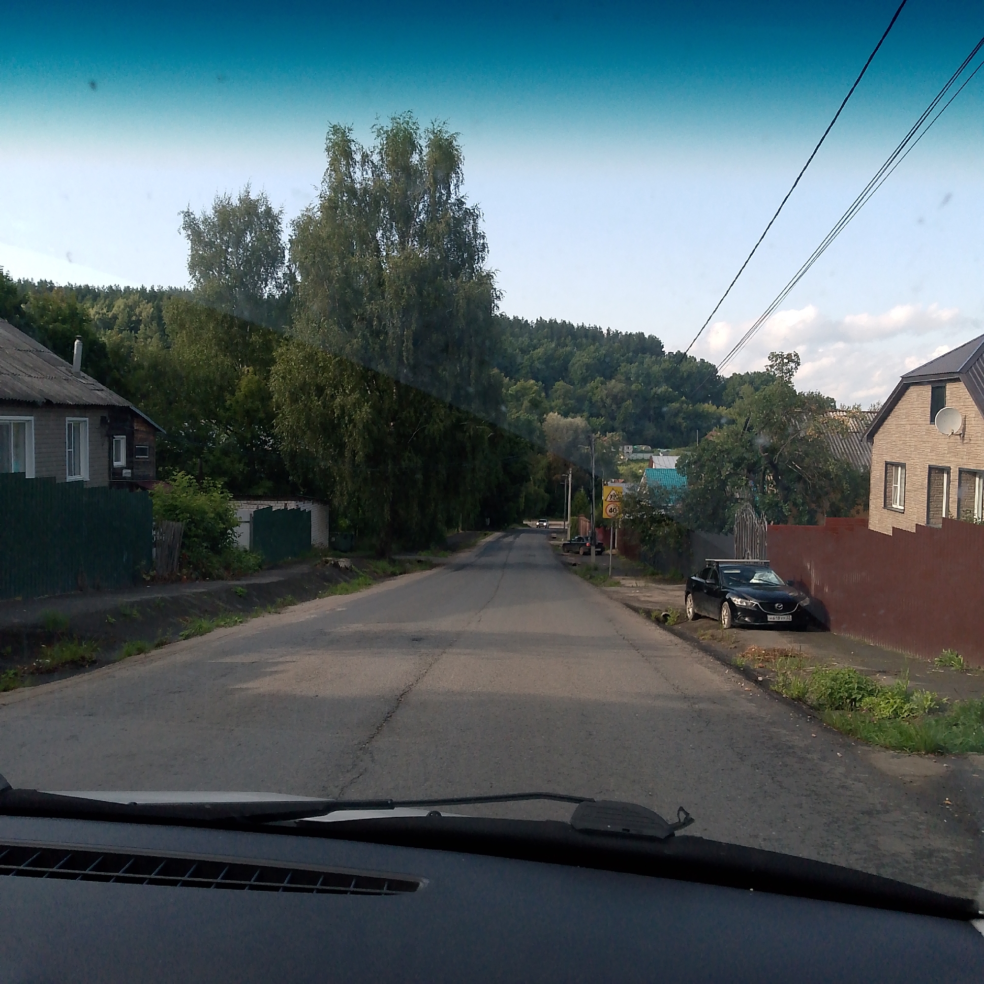 A view from inside a vehicle traveling on a paved road in a rural or suburban area of Vyazniki, Russia. The road is flanked by residential buildings, fences, and mature trees. On the right side of the road, a dark-colored sedan is parked near a brown metal fence. A speed limit sign indicating 40 km/h is visible near the parked car. The sky is a gradient of blue, with some clouds visible. The foreground shows the interior of the vehicle, including the dashboard and windshield.