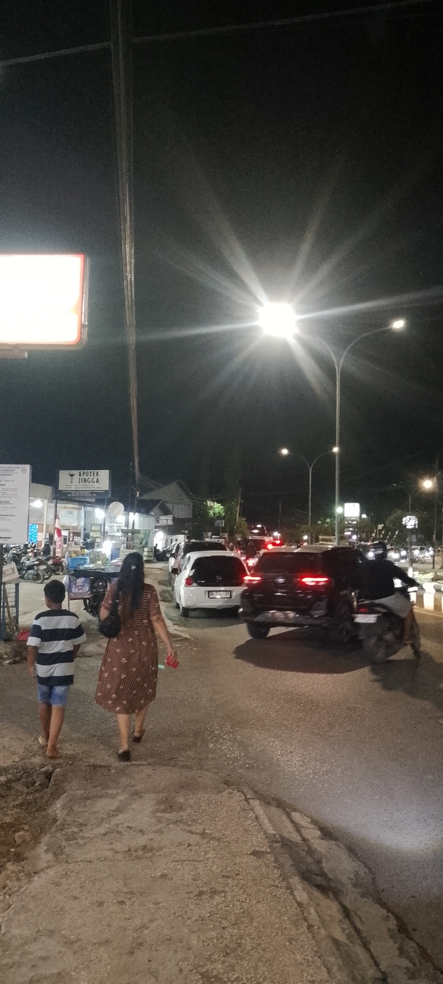 The image shows a street scene at night in Bersujud, Indonesia. A woman and a boy are walking away from the camera on a paved path. The woman is wearing a brown patterned dress and the boy is wearing a striped t-shirt and blue shorts. They are walking towards a street with traffic. Several cars are visible on the street, including a white hatchback and a black SUV. Streetlights illuminate the scene, with one particularly bright streetlight creating a starburst effect. In the background, there are buildings, including one with a sign that reads "APOTEK JINGGA". Motorcycles are parked on the side of the road near the buildings. The overall environment appears to be a commercial or residential area.