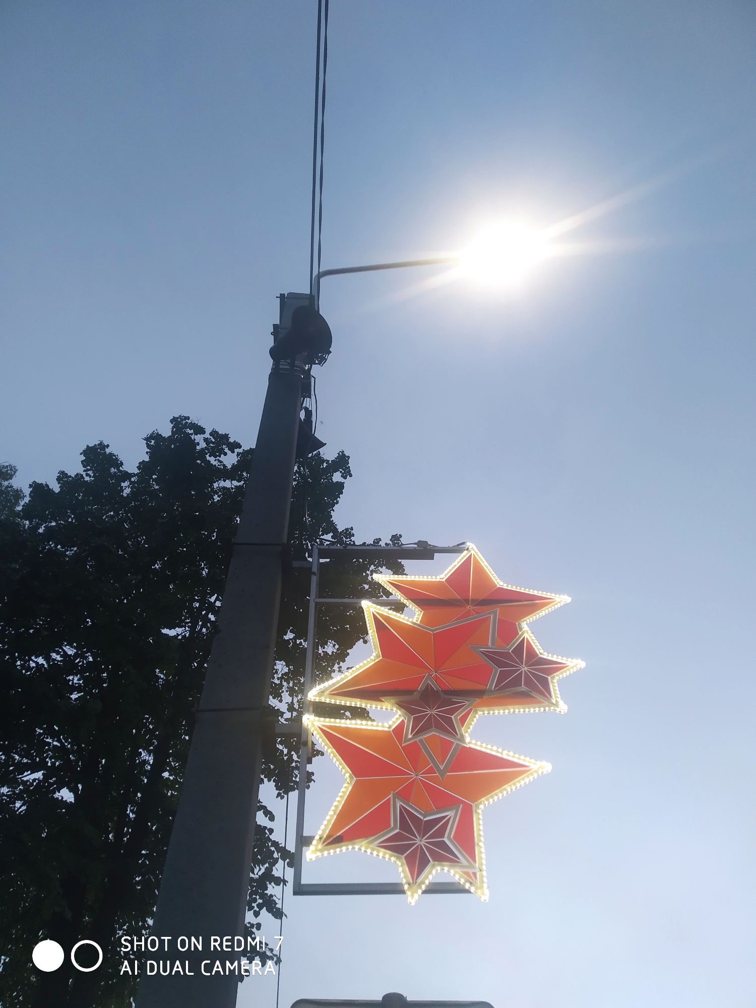The image shows a light pole in Chaikovsky, Russia, adorned with three red stars. The stars are arranged vertically, with the largest at the bottom and the smallest at the top. The stars are outlined with lights. A street light is attached to the pole above the stars. A tree with green foliage is partially visible to the left of the pole. The sky is light blue. The words "SHOT ON REDMI 7 AI DUAL CAMERA" are at the bottom of the image.