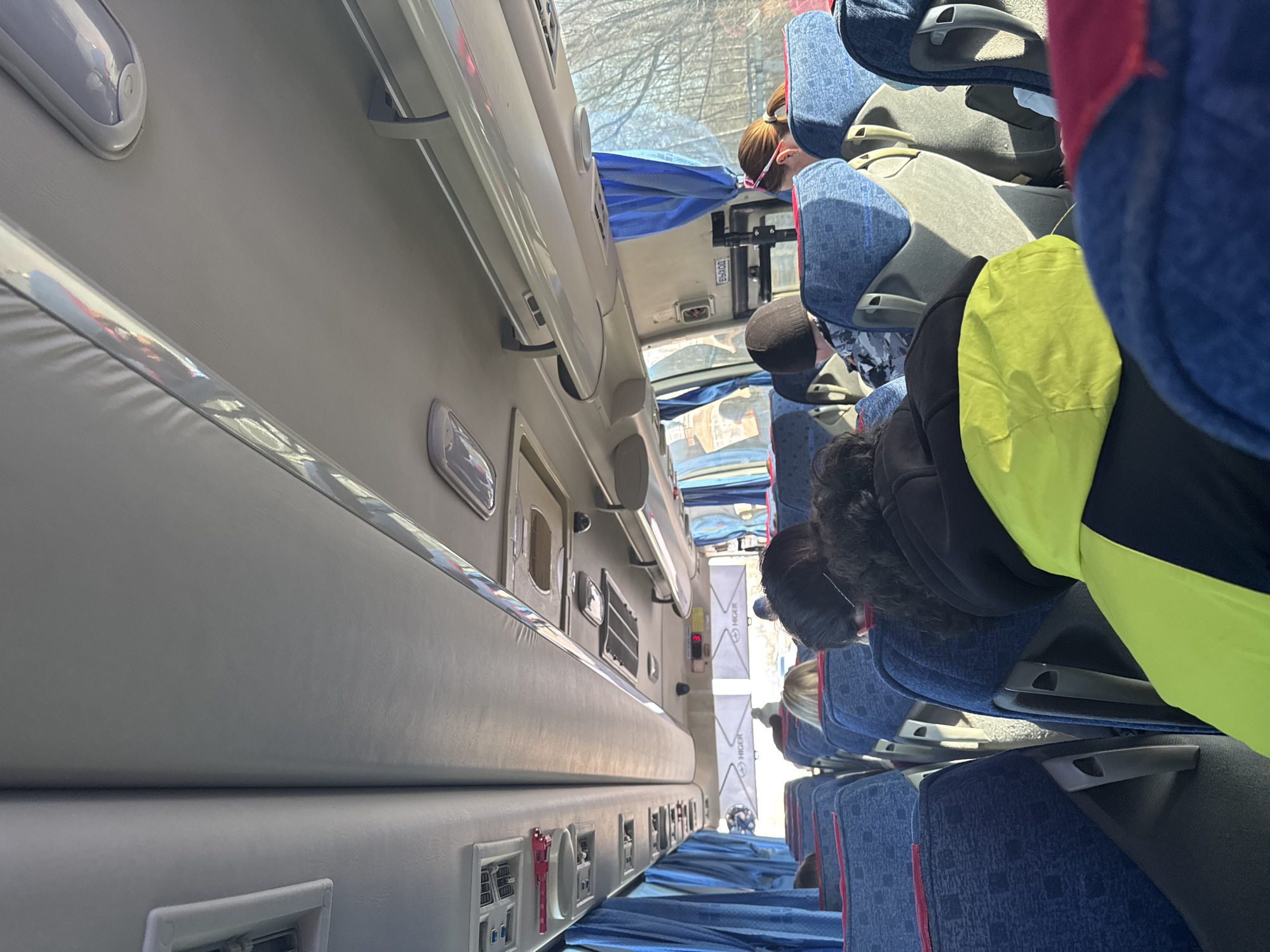 The image depicts the interior of a bus. Seated passengers occupy blue and red patterned seats. The bus interior includes overhead lighting, and a panel with charging ports and controls. A visible sign displays the word "HIGER". The location in the city of Berezniki, Russia cannot be determined from the image.