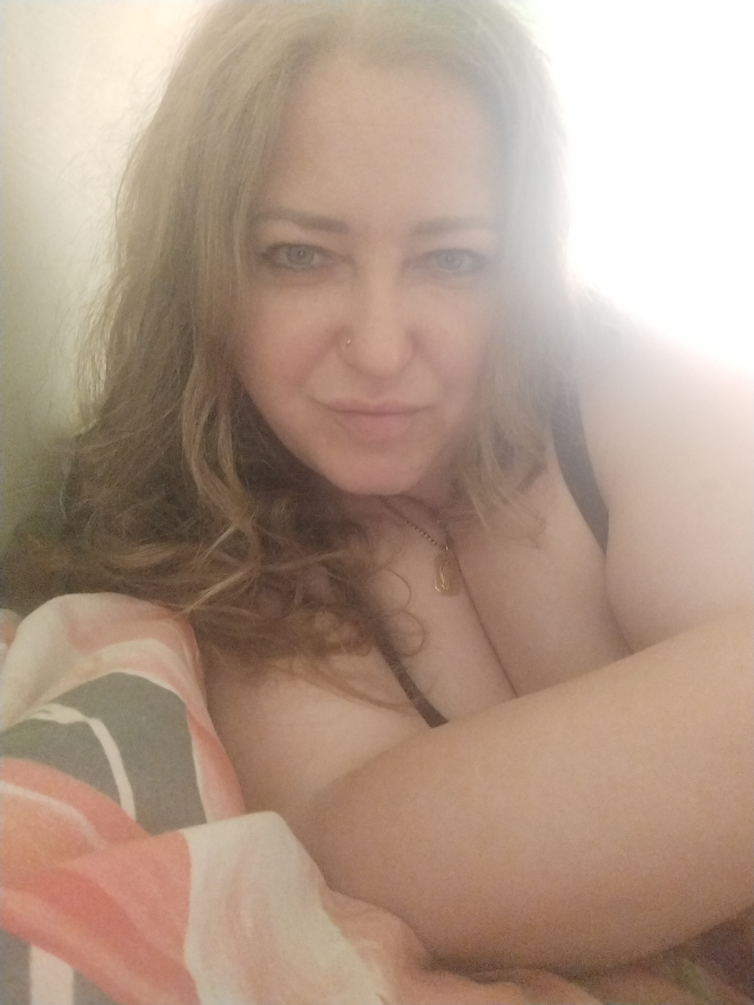 A woman is lying down in bed, facing the camera, with her head slightly tilted. She has long, wavy blonde hair and is wearing a necklace with a pendant. Her eyes are blue, and she has a small nose stud. The bed is covered with a duvet that has a pattern of pink, white, and gray stripes. The lighting in the room is bright, with a strong light source coming from the right side of the frame, creating a slight overexposure.