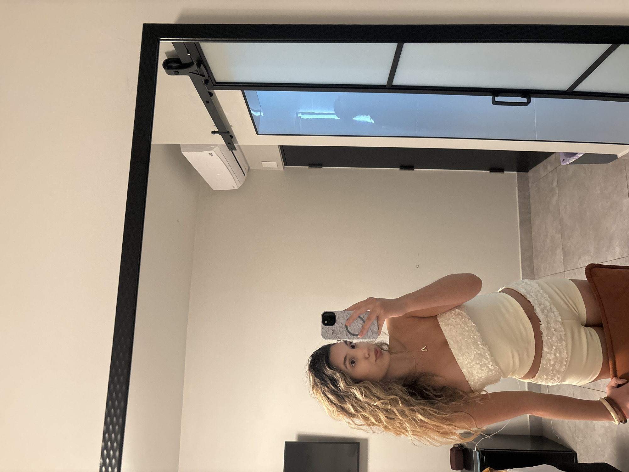 A young woman with long, curly blonde hair is taking a mirror selfie in a room. She is wearing a white strapless top with sequined detailing and matching shorts. A gold necklace with the letter "A" is visible around her neck. Her left arm rests on a brown chair. In the background, a large black-framed mirror reflects the room. To the right of the mirror, a sliding glass door with frosted panels is visible, along with a black door. An air conditioning unit is mounted on the wall above the doors. The flooring appears to be tile. The overall impression is an indoor setting, likely a hotel room in San Juan, United States of America.