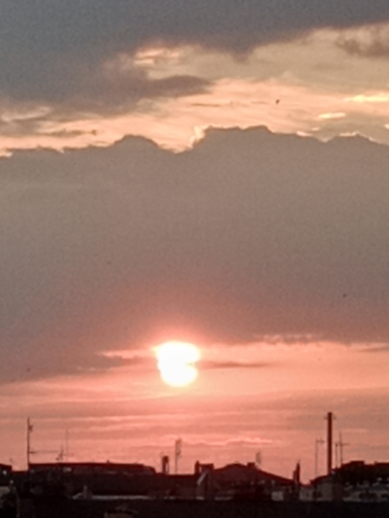 A blurry, low-resolution image depicts a sunset over a city skyline. The sun, a bright orb with a halo of light, is positioned in the lower-middle section of the frame, partially obscured by clouds. The sky is a gradient of orange and pink hues, with darker grey clouds forming a band across the top of the image. Silhouetted against the sky are the indistinct shapes of buildings, rooftops, and antenna-like structures, suggesting an urban environment. The overall impression is one of twilight or dusk, with the low light and soft focus contributing to a hazy atmosphere.