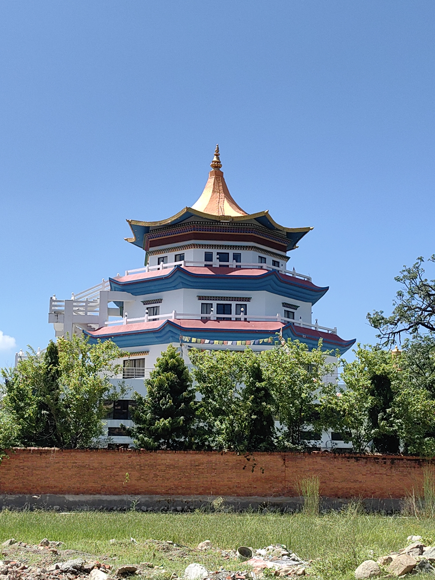 The image shows a multi-story building with a distinctive pagoda-style roof, adorned with gold, red, and blue accents, situated in Parsa, Nepal. The building is surrounded by lush green trees and is separated from the foreground by a brick wall. The sky is clear and blue. In the foreground, there is a patch of green grass with some scattered debris, suggesting an undeveloped or construction area.