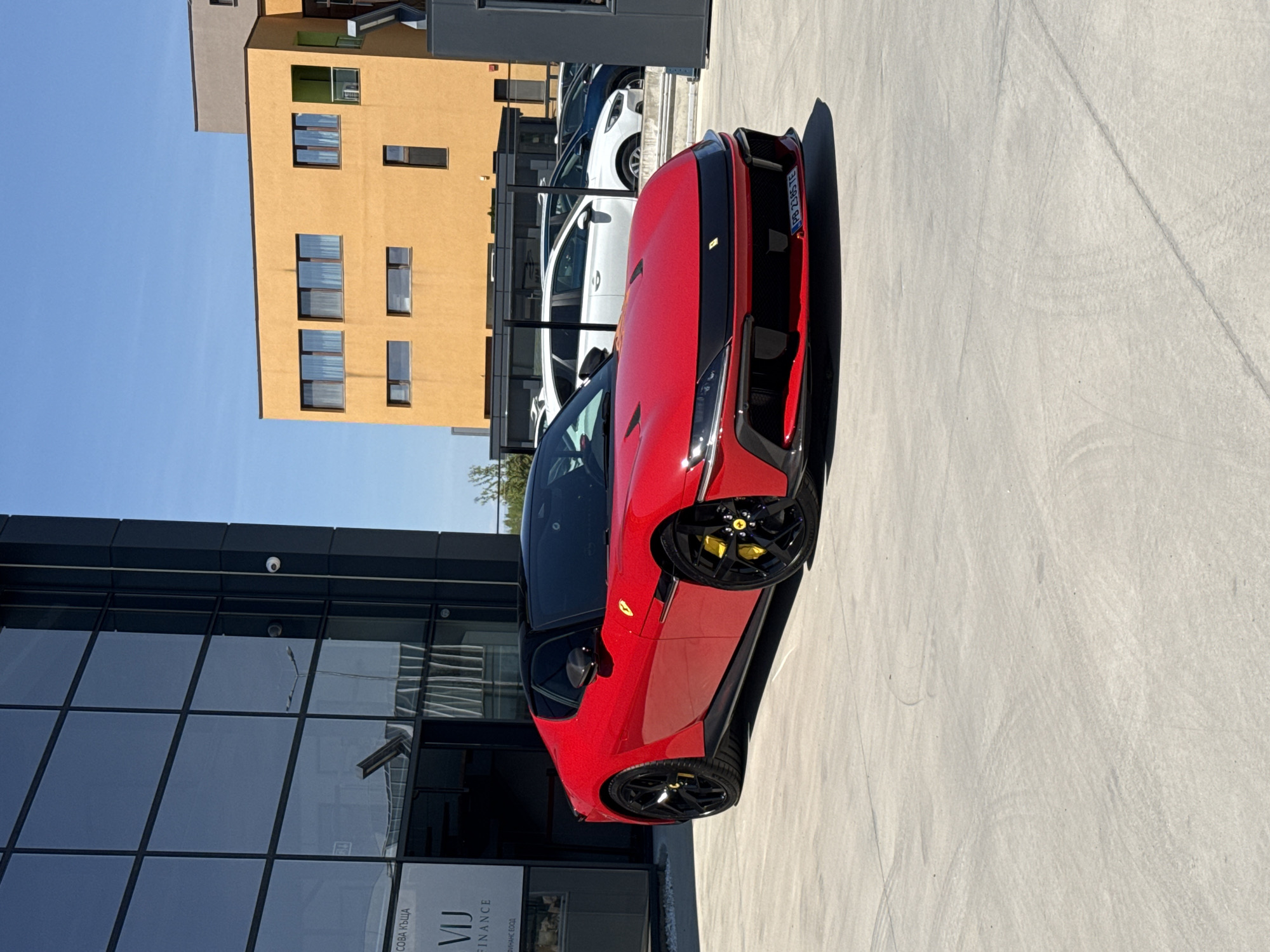 The image shows a red Ferrari parked in front of a modern building with a glass facade in Plovdiv, Bulgaria. The building features "VIJ FINANCE" signage in Cyrillic. Behind the Ferrari, there's an adjacent building with a yellow facade and numerous windows. Several cars are parked further in the background. The sky is clear and blue.