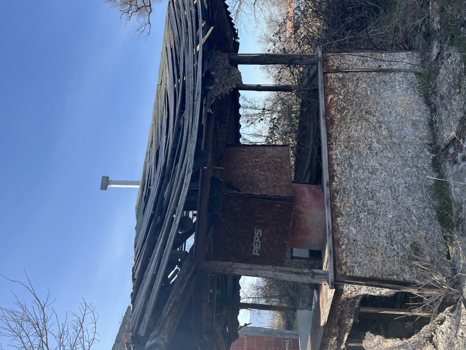 The image depicts a severely dilapidated, abandoned outdoor structure, likely a former kiosk or small commercial stall, located in Novakovo, Bulgaria, under a clear blue sky. The structure features a heavily damaged, sagging roof composed of multiple layers of dark, corrugated material, extensively torn and exposing underlying wooden rafters. The lower walls are constructed from weathered oriented strand board (OSB) or chipboard, with a significant section on the left exhibiting a large hole or collapse. Inside the structure, a brown panel bears the white letters "PEPS" (part of a "PEPSI" logo), affixed to an interior wall above a reddish, cuboid base. A slender, white cylindrical metal chimney with a rectangular cap rises vertically in the background, extending above the structure's roofline. The surrounding environment consists of overgrown, dry bushes and bare tree branches, indicating a neglected state. No individuals or active events are present in the scene.