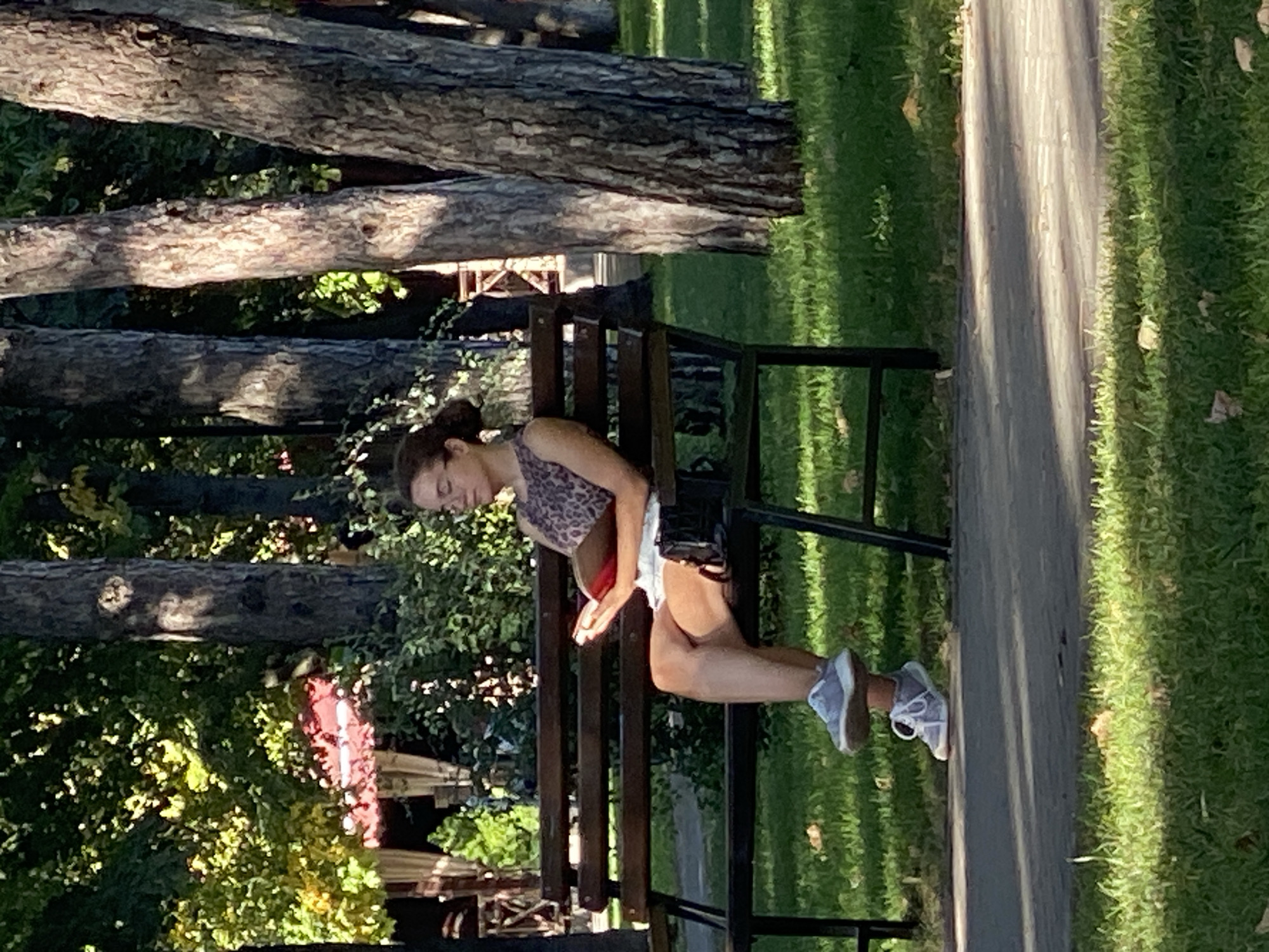 A woman wearing glasses and a leopard-print top with white shorts is sitting on a park bench reading a book. Her hair is pulled back in a bun. She is wearing grey sneakers. The bench is made of dark wood with metal legs. The bench is in a park with trees and green grass. The sun is shining, casting shadows on the ground. The image appears to be taken in Chișinău, Moldova, as suggested by the presence of mature trees often found in the city's parks and the overall atmosphere of a European park setting.