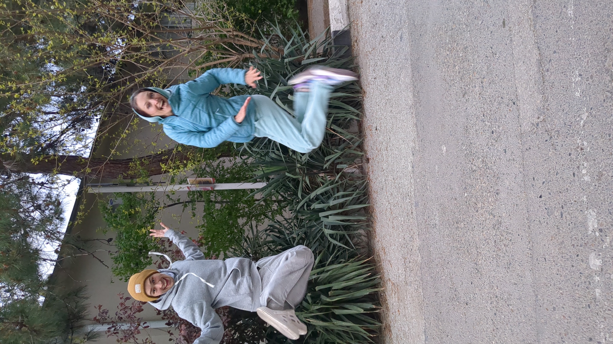 This image, set in Gelendzhik, Russia, captures two individuals in dynamic, mid-jump-like poses in an outdoor, park-like environment. The photo appears to have been taken with the camera rotated 90 degrees to the left, as the subjects and ground are oriented vertically.
The person at the top of the frame is wearing a light blue hooded sweatshirt and matching light blue sweatpants, with multicolored athletic shoes. They are captured mid-air, smiling broadly with their arms bent and hands open, conveying a sense of joy and movement.
The second person, located at the bottom of the frame, is dressed in a grey hooded sweatshirt, grey sweatpants, a yellow beanie, and light-colored slip-on shoes or clogs. They are also in a playful, airborne pose, with arms outstretched and knees bent, displaying a cheerful smile.
The background consists of various trees, some with delicate green leaves indicating springtime, and denser evergreen foliage. A light-colored building is partially visible through the branches. A dense strip of spiky green plants separates the subjects from the more distant trees and building. A light grey concrete or asphalt path, textured with small aggregate, runs along the right side of the frame, forming the ground surface. The lighting is soft and even, suggesting an overcast day or indirect sunlight. Both subjects clearly exhibit happiness and playfulness. No visible text is present in the image.