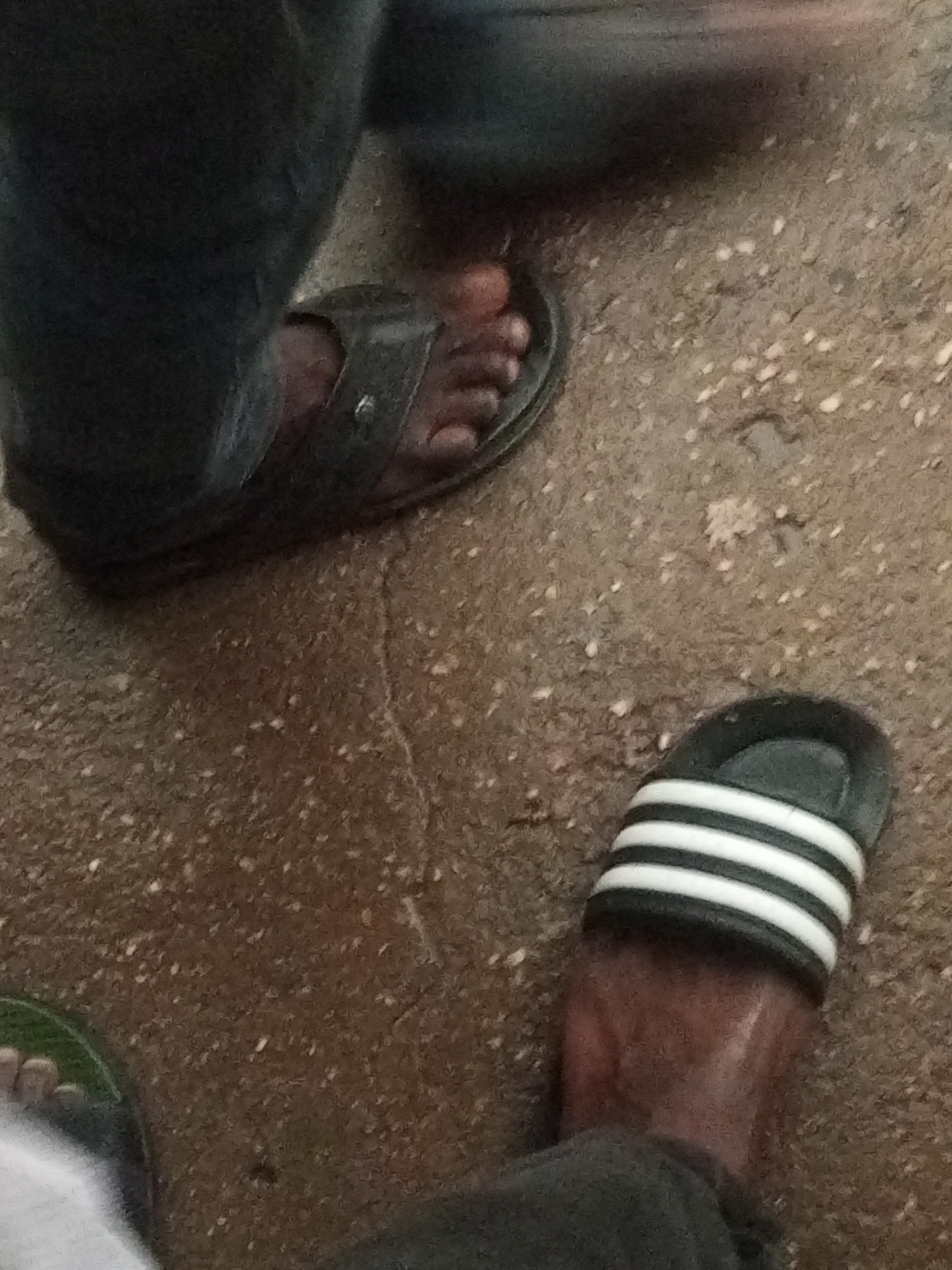 The image captures a low-angle perspective of the feet of at least two individuals, standing or moving on a speckled, brownish-grey ground surface, likely concrete or pavement, in Maiburiji, Nigeria.
In the foreground and to the right, a dark-skinned foot wears a black slide sandal featuring three prominent white stripes, characteristic of the Adidas brand. To the upper left, another foot, also dark-skinned, is partially visible wearing a dark, open-toed sandal or slipper. This person appears to be wearing dark trousers or pants. A third foot, wearing a green and white sandal, is partially visible in the bottom-left corner of the frame.
The ground appears slightly damp or wet, contributing to its textured, speckled appearance. The lighting is somewhat dim, suggesting an indoor setting, an overcast day, or shaded area. There is some motion blur, particularly in the upper right quadrant, indicating movement within the scene. No explicit text is clearly visible on any of the sandals, though the design on the black slide is distinctly recognizable as Adidas. The image provides a candid, ground-level view of people's footwear and the immediate environment.