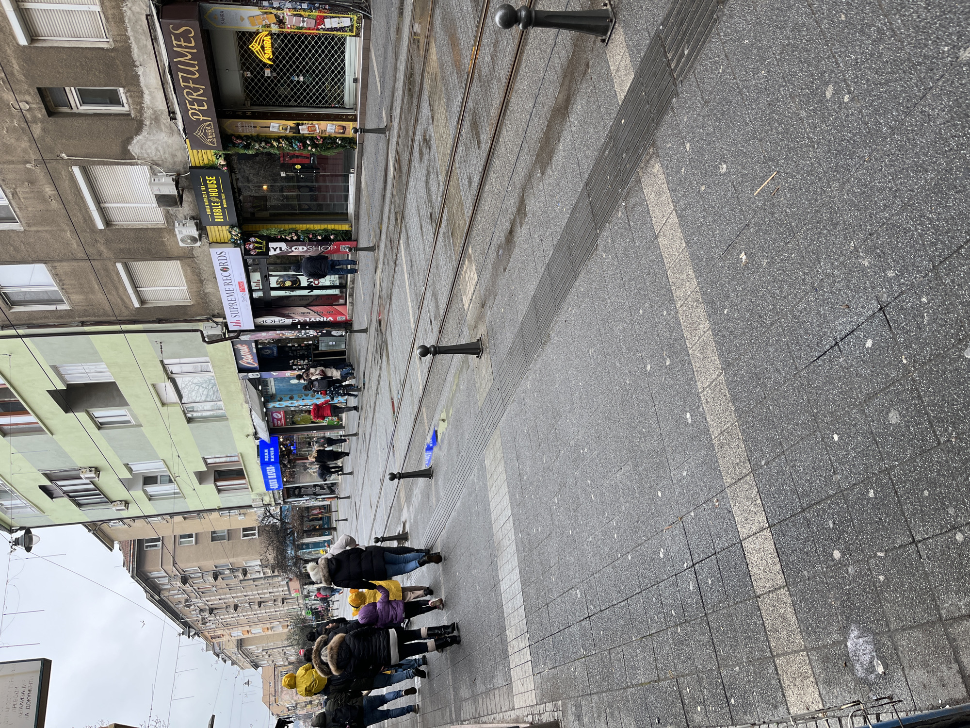 The image depicts a daytime street scene under an overcast sky, specifically Knyaz Boris I Street in Sofia, Bulgaria. The perspective is from a paved sidewalk looking down the street.
On the left, a group of approximately 15 pedestrians, predominantly adults, are walking away from the camera. This group includes at least two children, one wearing a bright yellow winter coat and another in a purple coat, accompanied by adults dressed in dark winter jackets and coats, some with fur-lined hoods. The sidewalk is paved with grey and lighter grey square tiles, featuring a tactile paving strip.
The center of the scene is occupied by two sets of wet tram tracks running parallel down the street. Dark, ball-topped bollards delineate the sidewalk from the tram track area and are also spaced along the tracks. A crumpled blue wrapper or piece of plastic is visible on the pavement near the tram tracks on the left side.
To the right of the tram tracks, a row of multi-story buildings lines the street. The closest building features a light green facade with horizontal white stripes. These buildings house various ground-floor retail shops. Visible shop signs include "SUPREME RECORDS" with a yellow awning, "BUBBLE HOUSE" (offering bubble waffles & tea) with a yellow facade and an entrance decorated with greenery, and "SAHRA PERFUMES" with a brown sign, its entrance secured by a metal roll-down shutter and featuring a "SAHRA" neon sign. Other shops display signs with Cyrillic script. Air conditioning units are mounted on the building facades. Scattered individuals are walking on the pavement in front of these shops.
Overhead, utility lines and streetlights traverse the street. The overall environment is urban, indicating a commercial and residential district.