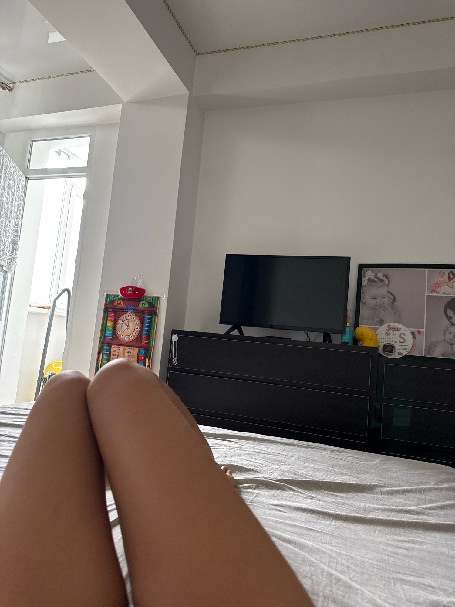 The image depicts a person's legs resting on a bed in what appears to be a bedroom. The room is illuminated by natural light coming from a window on the left, partially covered by a patterned curtain. To the right of the window, a large black dresser is visible, topped with a television, a framed collage of photographs, a yellow toy, and some small bottles. A colorful learning clock is hanging on the wall next to the dresser. The overall impression is one of a domestic setting, likely a bedroom, with personal items and decor suggesting a lived-in space. The image was captured in Chișinău, Moldova.