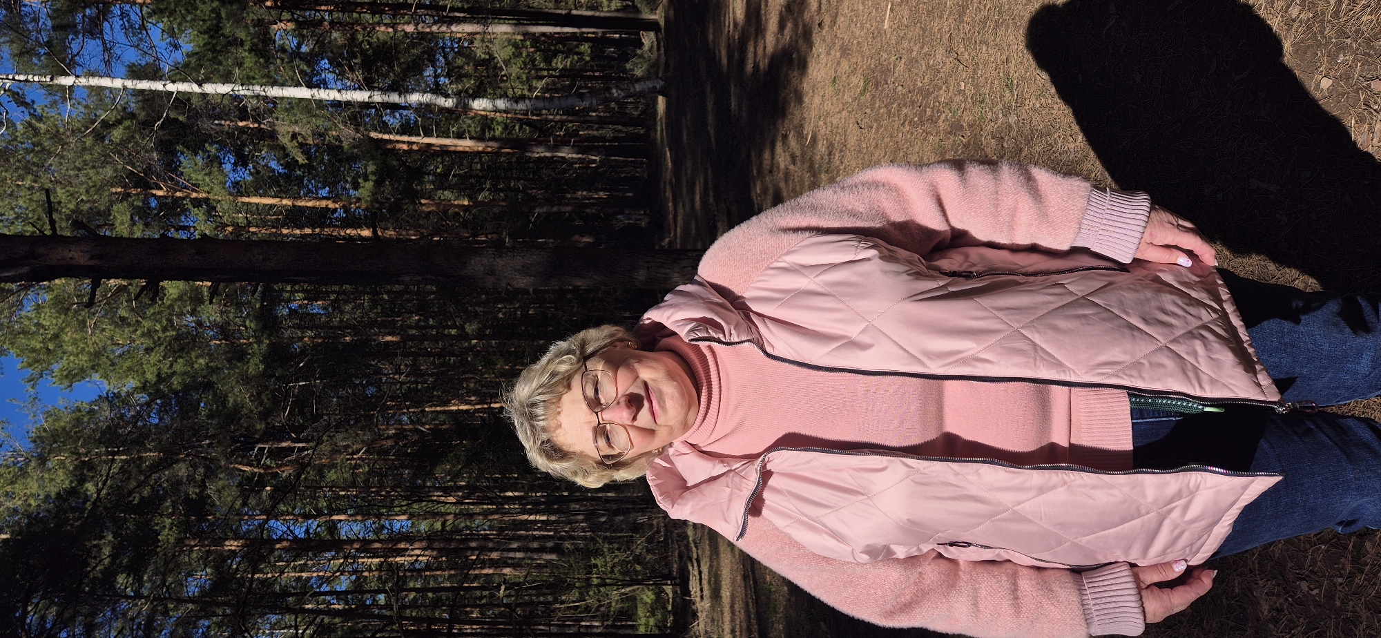 An older woman with blonde hair and glasses is captured in a forest setting, looking towards the camera with a gentle, pleasant expression. She is wearing a light pink turtleneck sweater, a matching light pink quilted vest with dark zippers, and blue jeans. Her hands, with light-colored nail polish, are resting in front of her.
The background consists of numerous tall pine trees with dark trunks and green needles, along with at least one birch tree identifiable by its white bark. Patches of clear blue sky are visible through the dense canopy of the trees. The ground is covered with dry brown leaves, dirt, and some sparse dry grass. Strong, dark shadows are cast on the ground to the right of the woman, indicating bright sunshine, likely in the late morning or afternoon. The overall impression is a sunny day in a natural, wooded environment. No visible text is present.