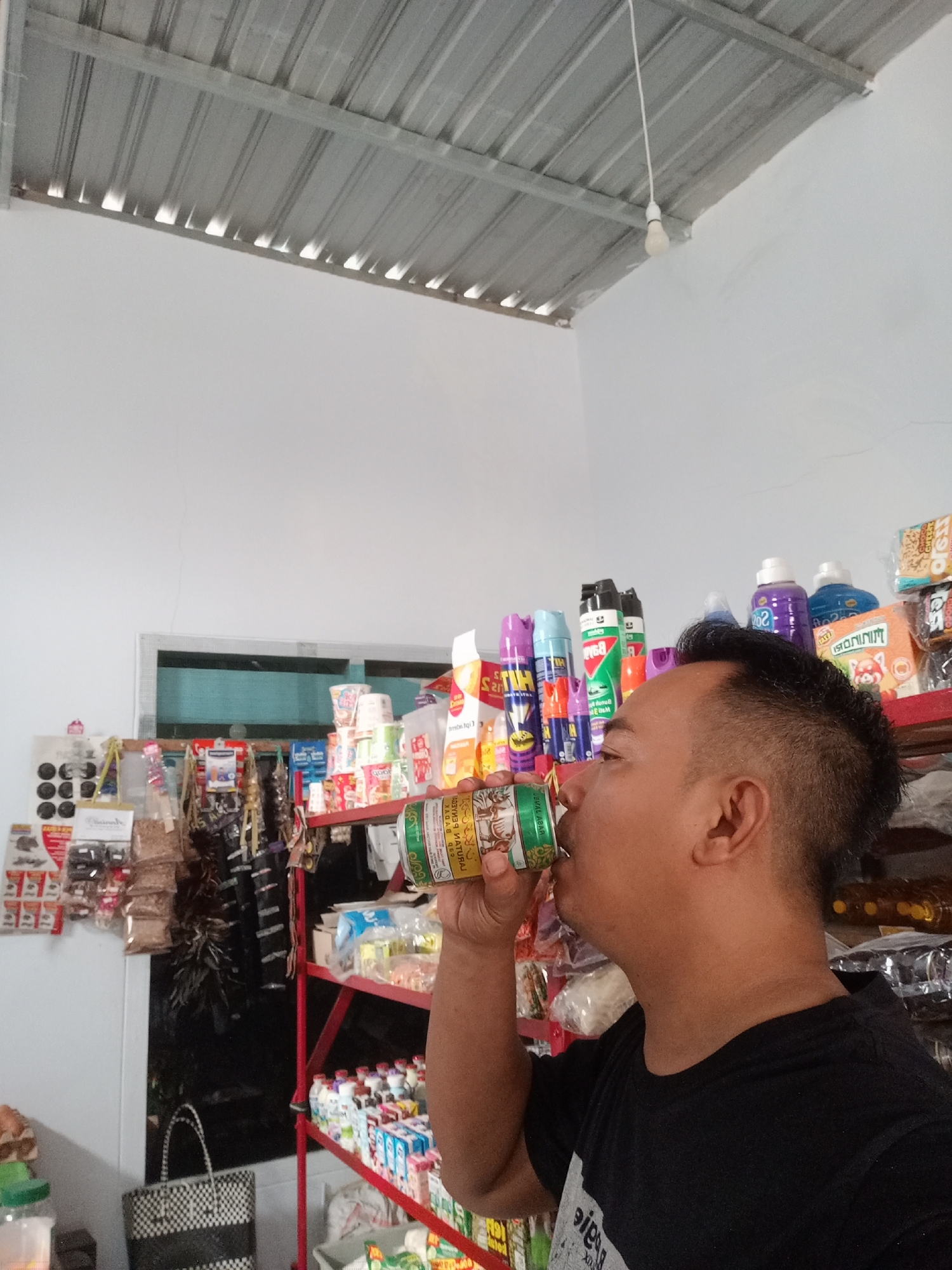 A man is drinking from a can in a small convenience store located in Simo, Indonesia. The man, seen from the side, is wearing a black t-shirt and has dark, spiky hair. The store is filled with shelves stocked with various products, including cans of drinks, cleaning supplies, and packaged goods. The ceiling of the store is made of corrugated metal, and a single light bulb hangs from the ceiling. The overall impression is that of a typical neighborhood store.