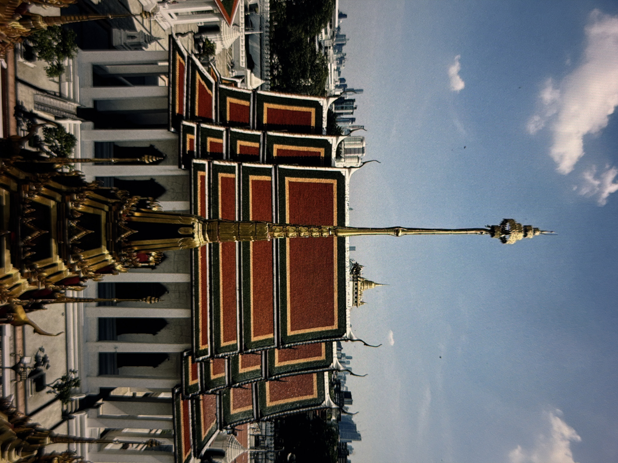 The image shows a building complex with tiered roofs and a golden spire. The roofs are red with gold and dark green trim. White pillars are visible along the building's lower level. In the background, a cityscape is seen against a blue sky with scattered clouds. The composition of the image suggests the building may be a temple or palace. The location is not in Da Nang, Vietnam. The image depicts Loha Prasat at Wat Ratchanatdaram in Bangkok.