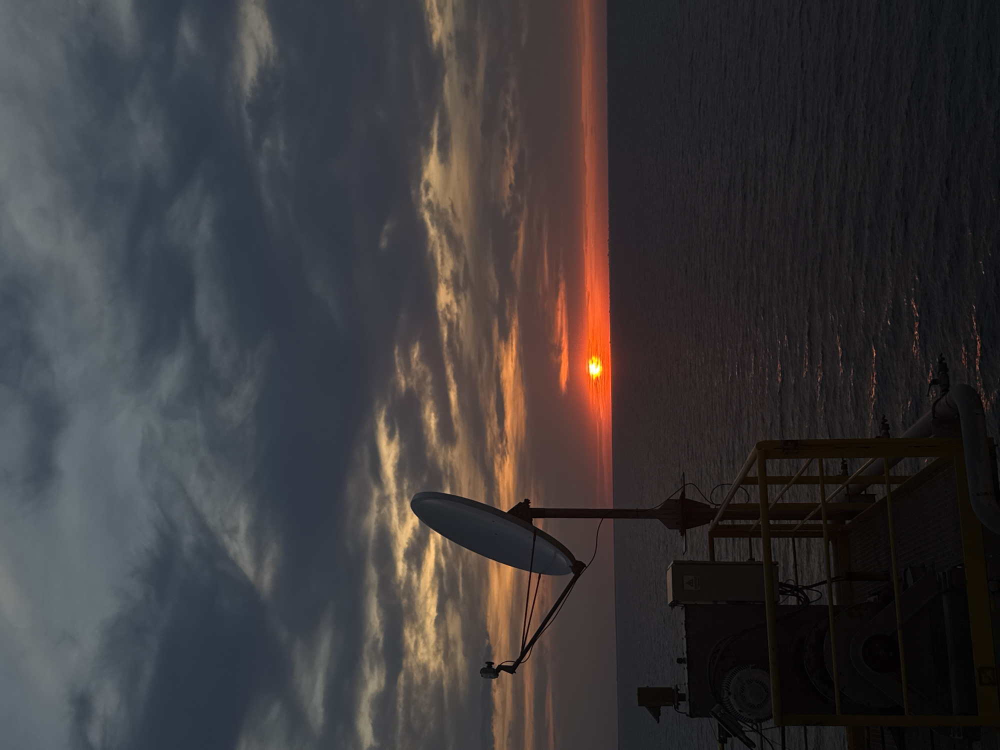 This is a photograph of a sunset over the ocean, viewed from what appears to be an offshore platform or ship. The sun is low on the horizon, casting a warm orange glow across the water and the underside of a cloudy sky. In the foreground, a satellite dish is mounted on a pole, silhouetted against the bright sky. To the left of the satellite dish, some industrial equipment is visible, including a large motor and various electrical boxes, all part of a yellow railing system. The sea is dark and appears choppy, with a few reflections of the sunset visible on its surface. The overall impression is one of a functional, industrial setting juxtaposed with the natural beauty of a sunset. The specific location is not identifiable within the image.