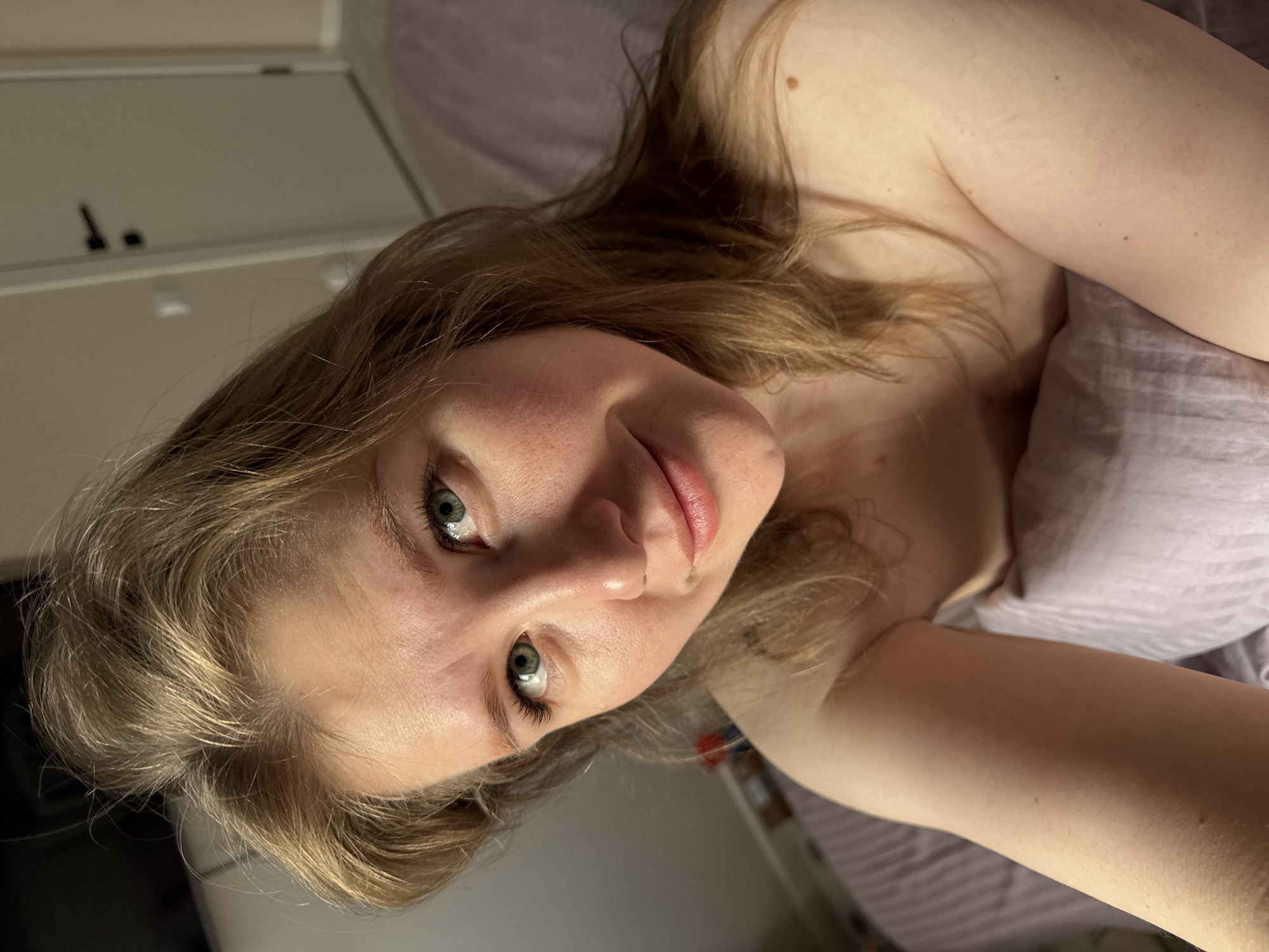 A woman with light brown hair and fair skin is lying in bed, covered with a lilac-colored duvet. She is looking directly at the camera. The background features a light-colored dresser with dark handles. The location is potentially within the city of Ферма, Russia, but this cannot be verified without further information.