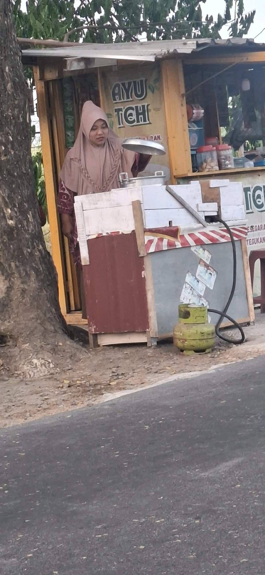 In Desa Gagaksipat, Indonesia, a woman wearing a light brown hijab and a patterned maroon dress stands behind a small, makeshift food stall. The stall appears to be constructed from wood and corrugated metal. A yellow banner with the words "AYU TCH" is visible on the stall. In front of her, she holds a metal tray. The stall is stocked with various items, including containers of food and a green propane tank connected to a hose. The scene is set outdoors, with a tree trunk visible to the left and a paved road in the foreground.