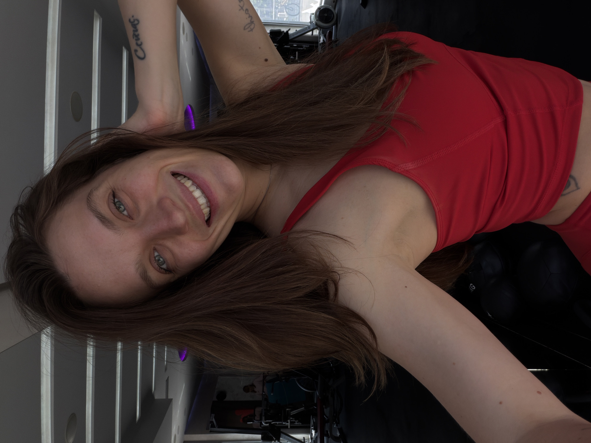 A woman with long brown hair, wearing a red athletic top, is pictured at a fitness center in Budva, Montenegro. She is smiling and positioned at an angle, revealing tattoos on her arms. Gym equipment is visible in the background, and the room has a modern design with light-colored walls.