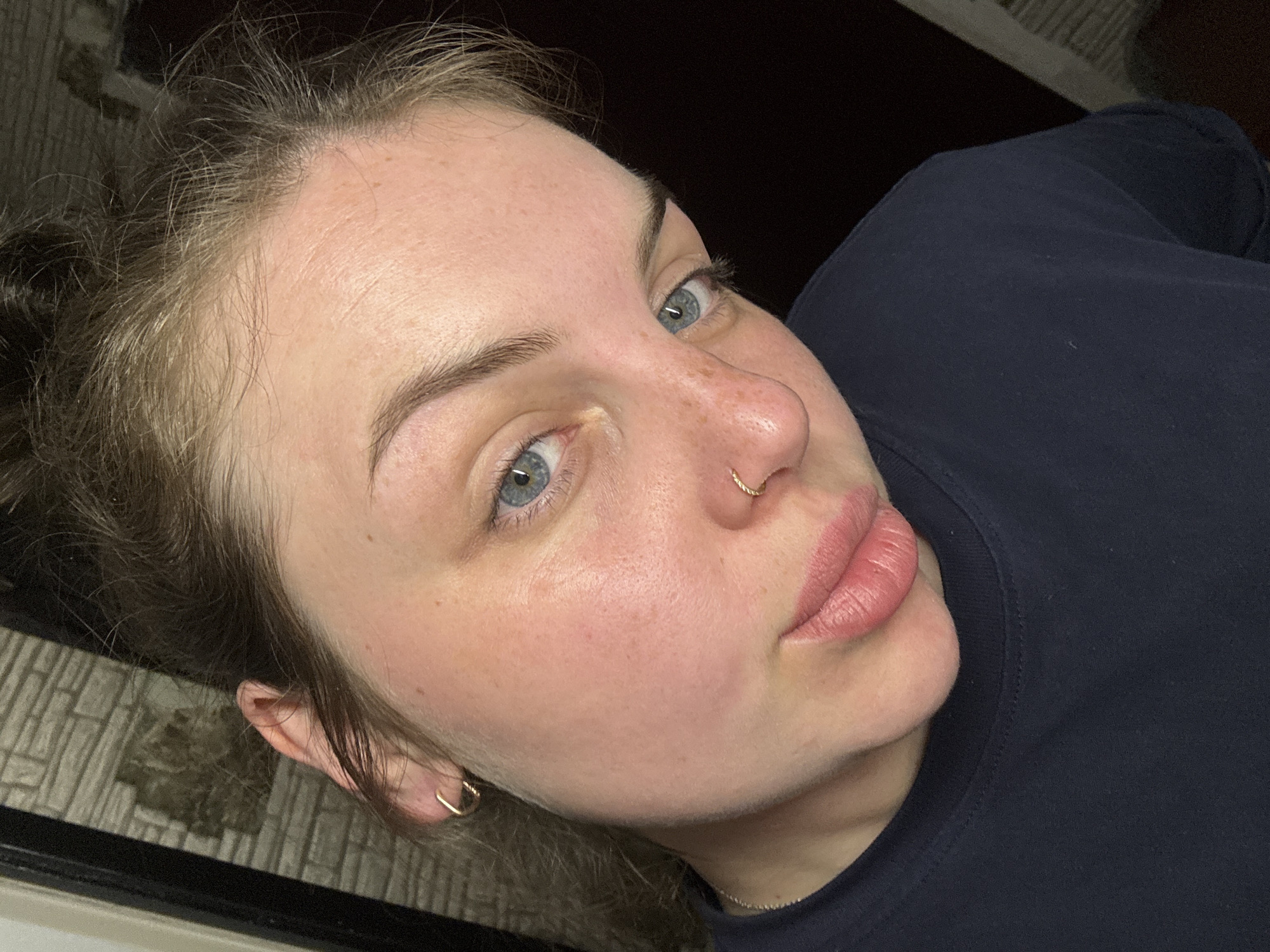 A close-up, selfie-style photograph shows a woman with light skin and light blue eyes. She has freckles across her nose and cheeks. Her hair is light brown, pulled back in a messy style. She is wearing a gold hoop earring in her left ear and a gold nose ring in her left nostril. She is wearing a dark blue t-shirt. The background is dark and out of focus. The photograph doesn't provide any information related to the city of Chornomorskoe, Ukraine.