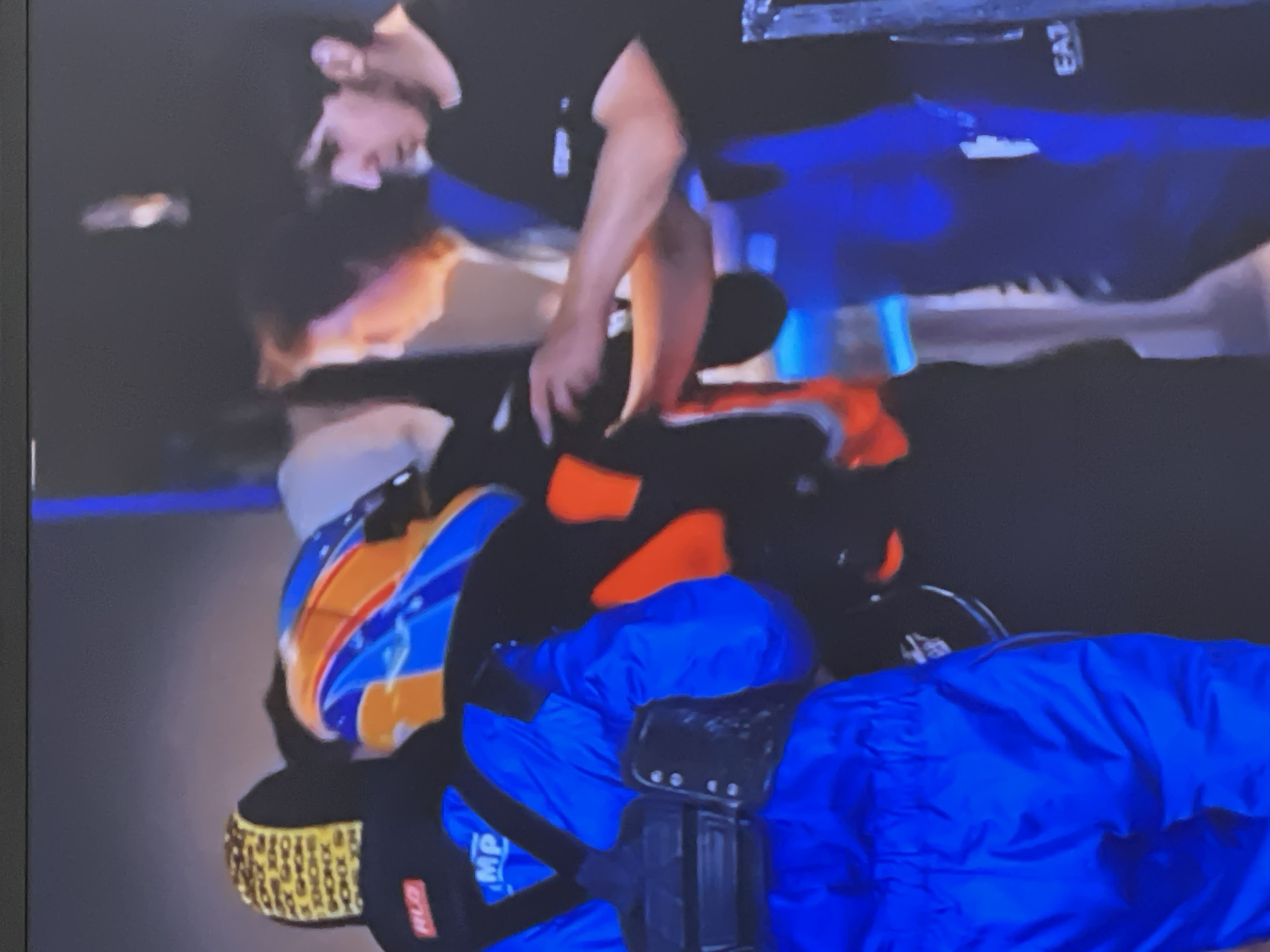 The image depicts a scene involving several individuals in an indoor or low-light environment, potentially in Moscow, Russia. In the central area, a person with dark hair and a beard, wearing a black shirt with "EA7" text, is bending over, appearing to assist another individual.
The person being assisted is wearing a distinctive helmet featuring blue, orange, and white/yellow patterns. They are also clad in an orange and black suit, possibly protective gear or a racing suit, with further blue elements visible on their lower body. A textured golden/yellow pattern is visible on the helmet or equipment of this individual.
In the foreground, another person is visible wearing a bright blue suit or uniform, with a black brace or harness fastened across their torso. Visible text on their clothing includes "MP" and "RLD". The background is dark and somewhat blurred, with hints of blue lighting or structures. The overall activity suggests a moment of practical assistance or preparation among individuals wearing specialized attire.