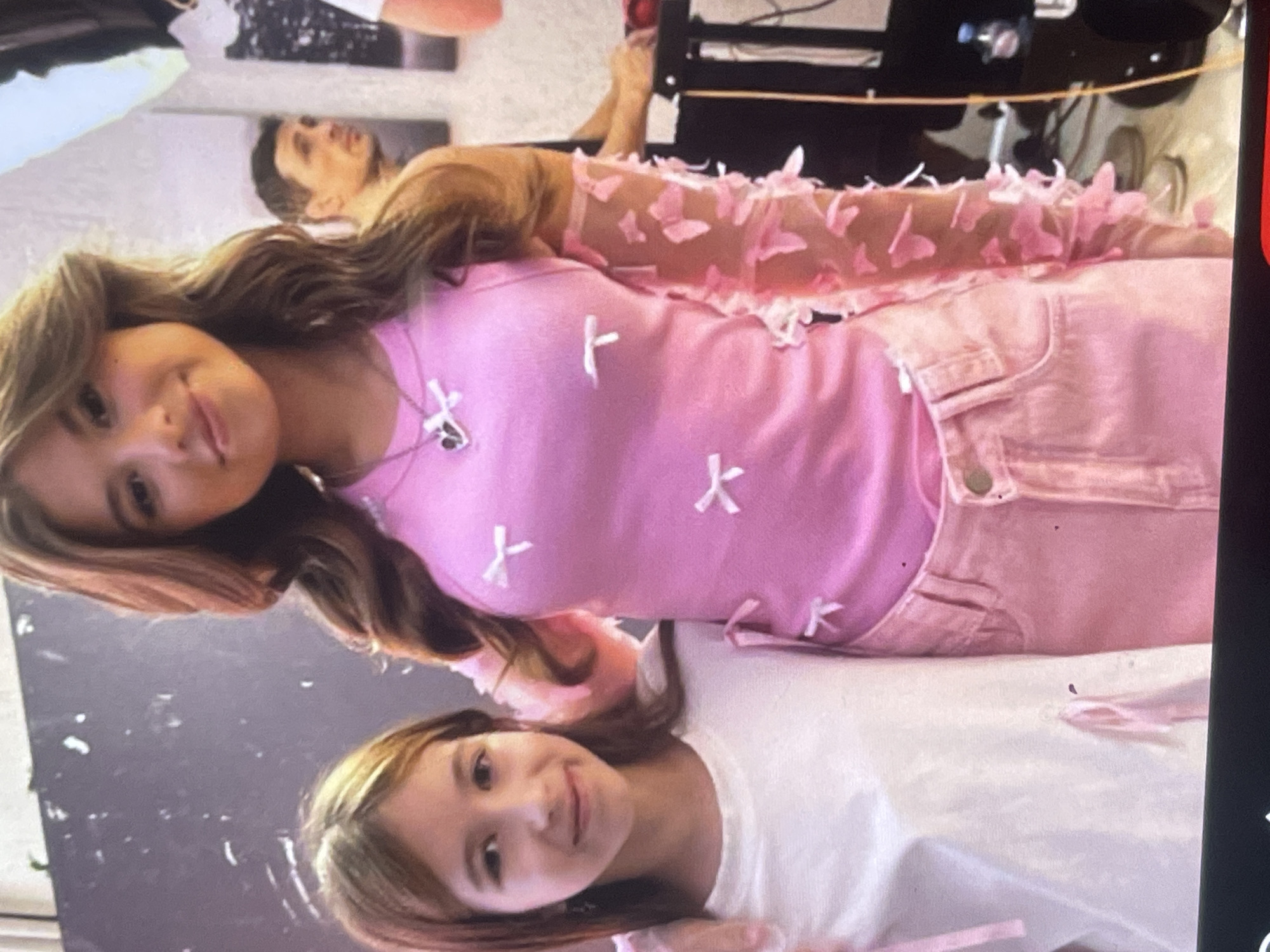 This photograph, likely taken indoors in Poltava, Ukraine, features a young woman as the primary subject, with a younger girl visible in a reflection and a man in the background. The young woman, with long, wavy light brown hair, is positioned centrally, looking directly at the camera with a gentle smile. She wears a vibrant pink short-sleeved top adorned with small white bow-shaped appliqués, paired with light pink denim-style pants or overalls. Her right arm is covered by a sheer, light pink sleeve decorated with numerous delicate butterfly cutouts. A silver-toned necklace with a small pendant is visible around her neck.
A prominent feature is the reflective surface in the foreground, showing a clear image of a younger girl, likely a child, with similar light brown hair. She is also looking towards the camera with a smile and is wearing a plain white t-shirt.
In the upper left background, an adult male with dark hair is partially visible and out of focus, looking towards the right. The indoor setting appears bright, possibly a studio or an event space, with dark, indistinct structures suggesting equipment or furniture in the deeper background. The overall activity involves the subjects posing for a photo, conveying a cheerful and relaxed mood. No discernible text is visible in the image.