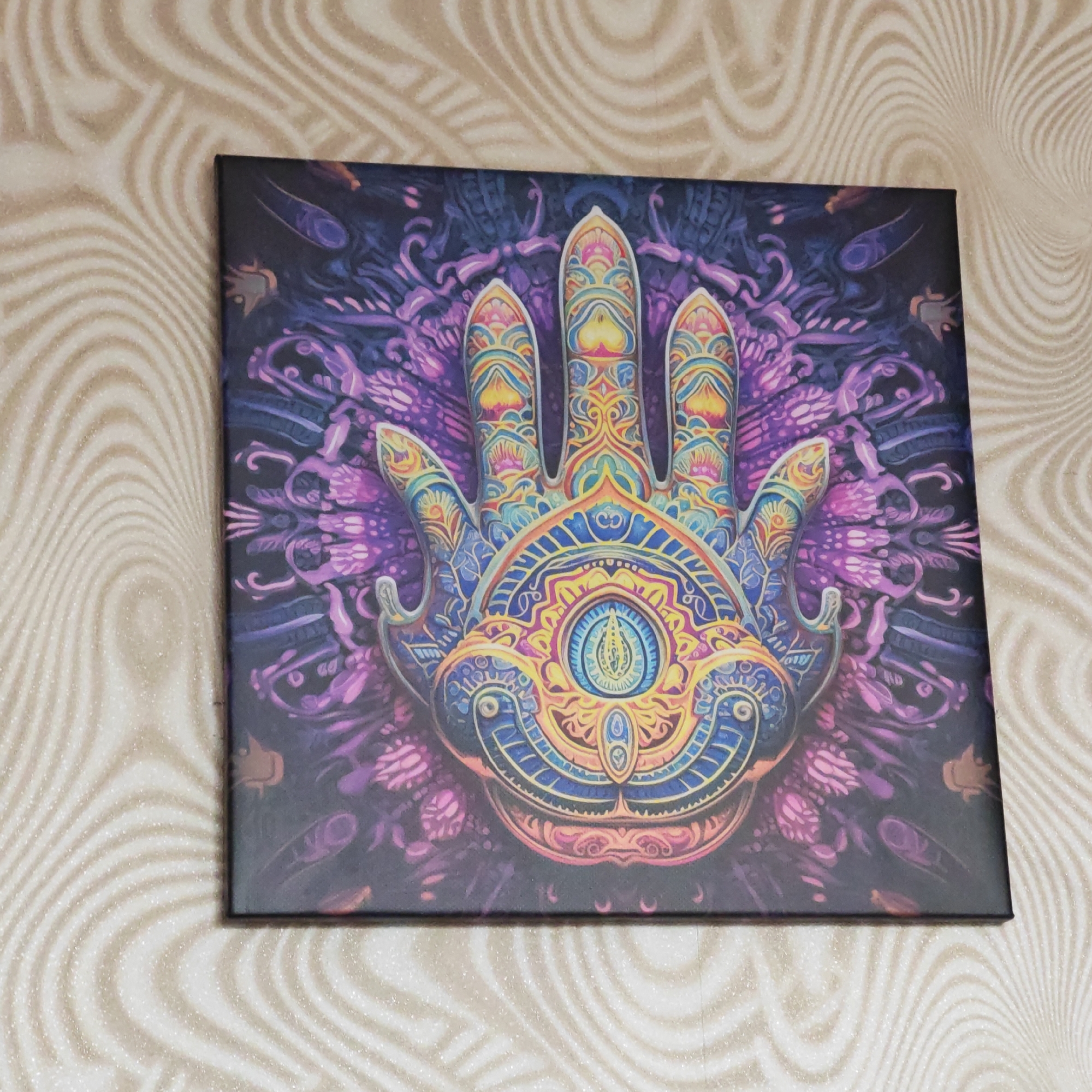 The image shows a square canvas print hanging on a wall with a wave-like pattern. The canvas features a vibrant, detailed illustration of a stylized hand with an eye in the palm, surrounded by intricate designs in shades of purple, blue, yellow, and orange. The environment is indoors. Location details such as the city or specific address cannot be determined from the image.