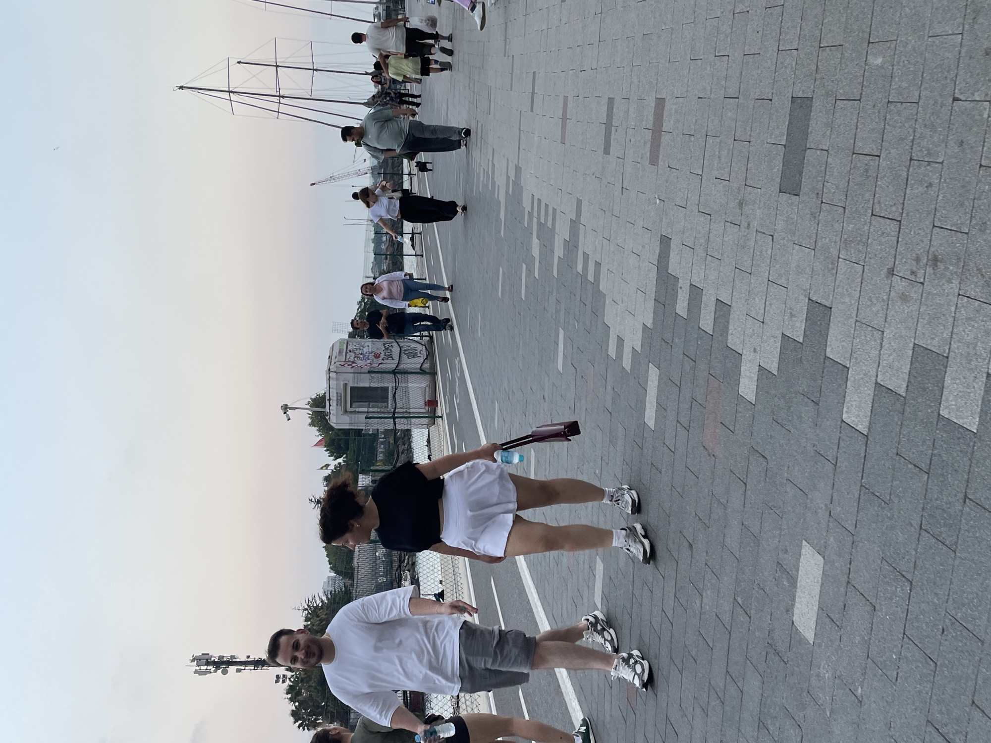 A street scene in Kadıköy, Turkey, depicts several individuals walking along a paved promenade by the water. In the foreground, a man in a white t-shirt and grey shorts walks with a woman in a black crop top and a white skirt. The woman carries a dark red purse and a water bottle. Further along the promenade, other people are seen walking, including a man in a grey t-shirt and dark pants, a child in a light green t-shirt, and a man in a white t-shirt. A small black dog is also visible on a leash. In the background, various boats and masts are docked, and a cell tower is prominent on the left side of the image. The sky is overcast, suggesting it is either early morning or late evening. The promenade is constructed with grey and light grey paving stones.