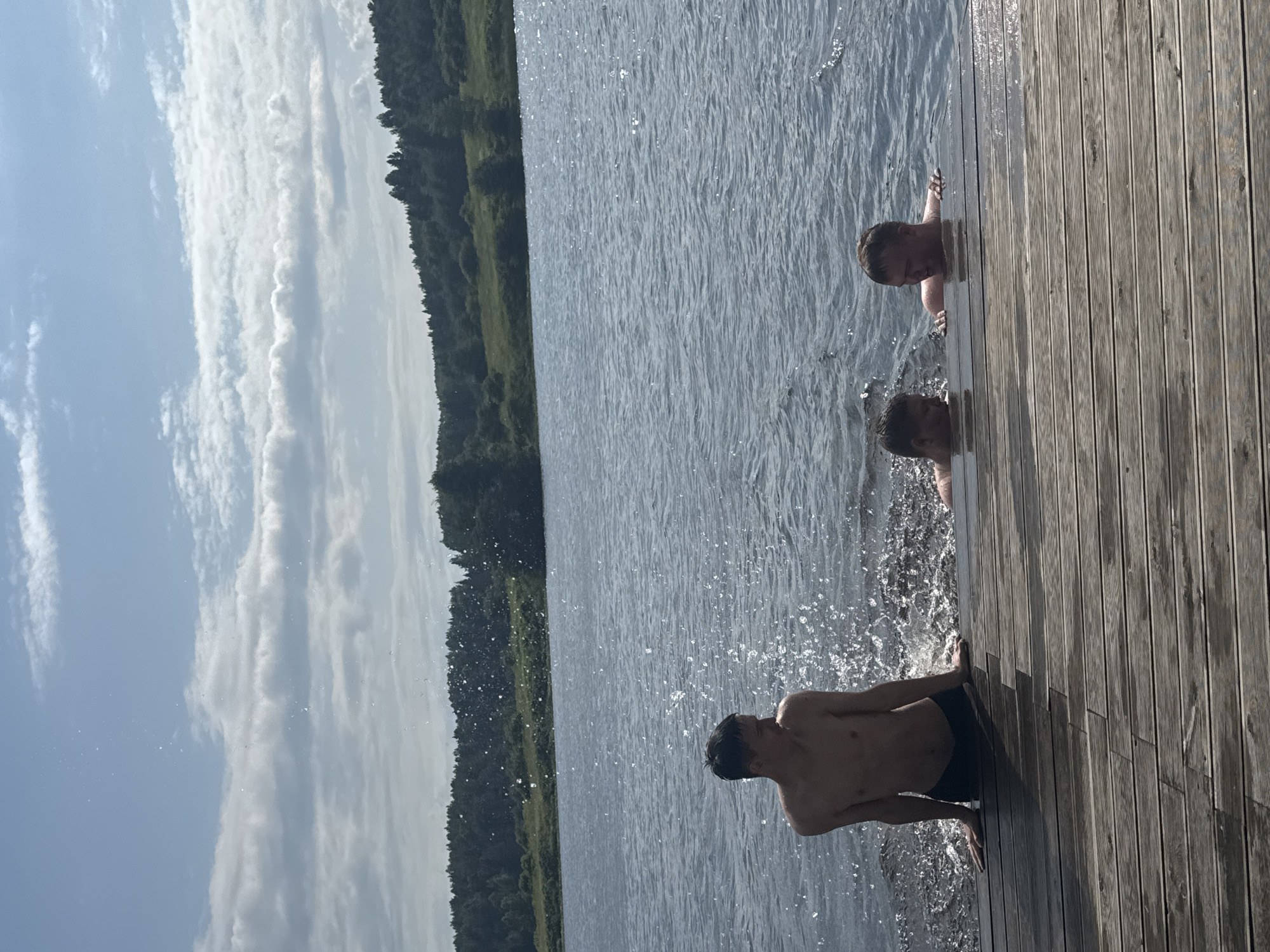 The image depicts three young men by a lake in Pozdyshki, Russia. Two individuals are in the water, with one holding onto a wooden dock and the other submerged with only his head visible. The third person is on the dock, wearing dark swim trunks and appearing to splash water. The scene is set on a sunny day, indicated by the bright sky filled with white, wispy clouds and the sunlit water. A dense forest lines the far bank of the lake, suggesting a natural, wooded environment. The wooden dock, made of weathered planks, runs along the right side of the frame, providing a clear vantage point to the water. The overall mood is one of leisure and enjoyment, with the subjects engaged in recreational activities on a warm day. There is no visible text in the image.