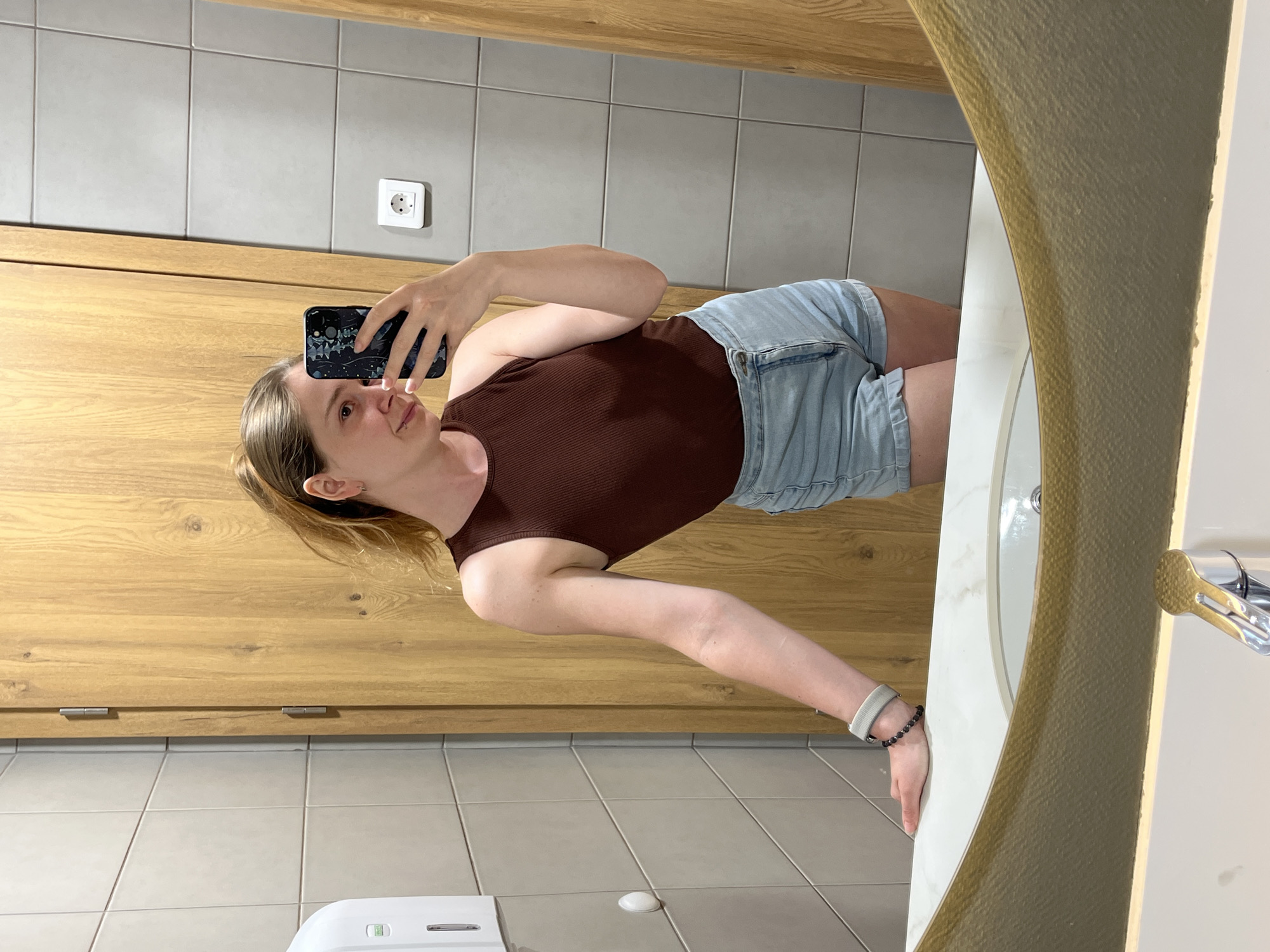 A young Caucasian woman is taking a mirror selfie in a modern restroom. She is wearing a brown ribbed tank top and light blue denim shorts. She has her hair pulled back in a ponytail and is wearing a bracelet on her left wrist. The restroom features light gray tiled walls, a wooden door, a white sink with a marble countertop, and a large mirror. A white soap dispenser is mounted on the wall to the left, and a white electrical outlet is visible on the right. The lighting appears to be artificial and ambient. The background also shows the corner of what appears to be a modern hotel or public building, possibly in the city of Plovdiv, Bulgaria, given the style of the tiles and woodwork.