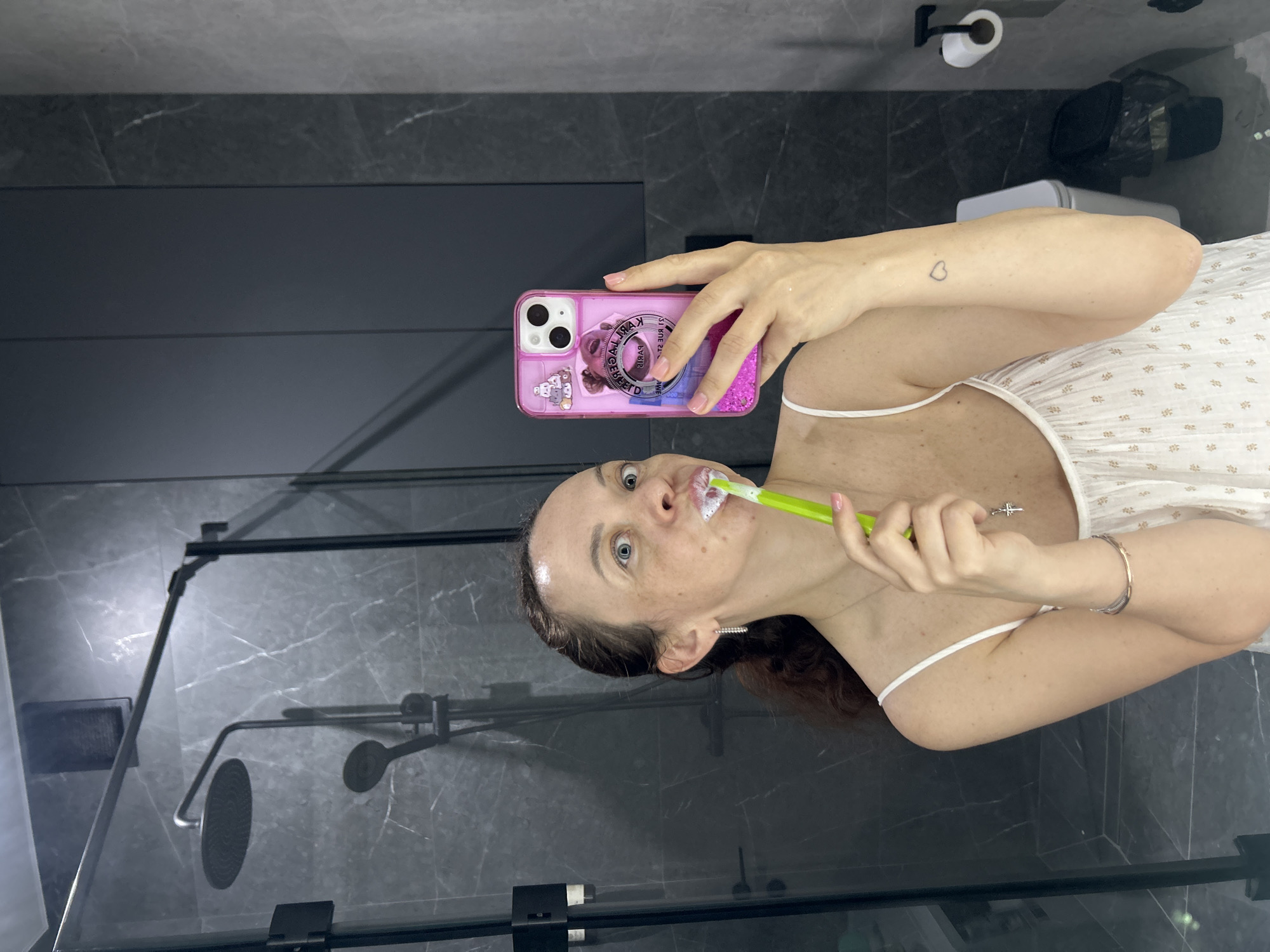 This image depicts a single woman engaged in a morning routine within a modern bathroom, captured as a mirror selfie in the city of Yekaterinburg, Russia.
The woman, with light skin and dark brown hair pulled back, is visible from the chest up, reflected in a mirror. Her eyes are wide, looking directly towards the smartphone she holds in her right hand. She is brushing her teeth with a bright green toothbrush, generating white foam around her mouth. She wears a white or light-colored camisole top with thin straps. Her left ear displays a small hoop earring, and a delicate necklace with a small cross pendant is visible on her chest. A multi-strand bracelet adorns her left wrist, and a small outline heart tattoo is visible on her right forearm.
Her right hand holds a light-colored smartphone, equipped with a translucent pink glittery case. The case features a circular pop socket with partially legible reversed text, including "PARIS," and a graphic that appears to include cute, possibly Sanrio-style, characters.
The bathroom environment is characterized by dark gray/black marble-patterned wall tiles with white veining. Behind the woman, a shower enclosure is visible with a round rainfall shower head and a handheld shower head, all in black. A glass shower door and its black supporting bar are also present. To the right, dark gray vertical-paneled cabinetry is integrated into the wall. Further to the right in the reflection, a white toilet with a closed lid, a black waste bin with a liner, and a black wall-mounted toilet paper holder with a roll of toilet paper are discernible. A black wall-mounted dispenser is also visible inside the shower enclosure. The overall aesthetic is clean and contemporary with a dark, monochromatic palette.