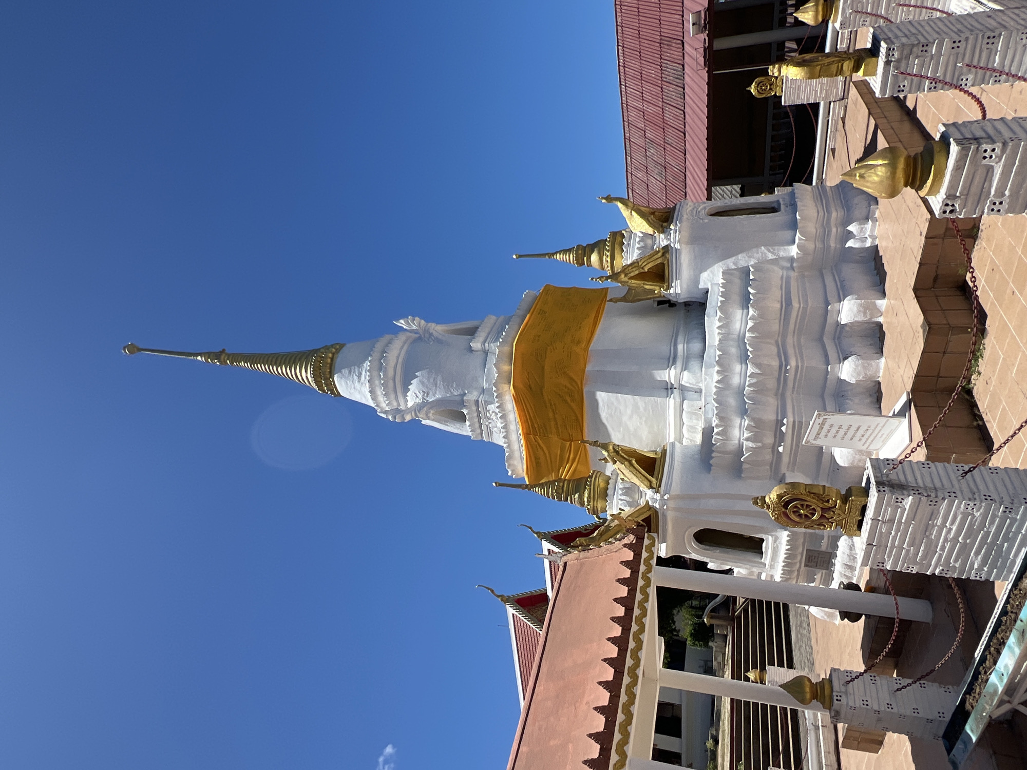 This media file shows a close-up of a white Buddhist stupa with a golden spire, wrapped in a bright yellow sash. The stupa is adorned with decorative golden accents, including figures resembling mythical creatures and what appears to be a Dharma wheel. The stupa is situated outdoors, with a clear blue sky visible. In the background, part of a building with a reddish-brown tiled roof can be seen. The architectural style suggests a Southeast Asian location, consistent with the provided context of Baan Hua Thanon, Thailand. The lighting indicates it is daytime, likely sunny, given the strong shadows and bright sky. There are no people visible in the frame. Some text is present on a white sign attached to the stupa, but it is too blurry to read. The overall scene depicts a religious structure in a well-maintained environment.